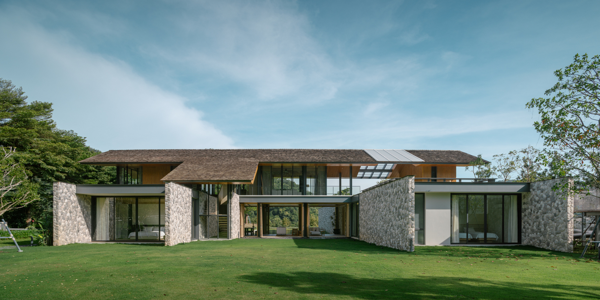 Front elevation of a low house with a shingled hipped roof, rubble stone walls and glazed ground floor, set above a lawn in clear daylight