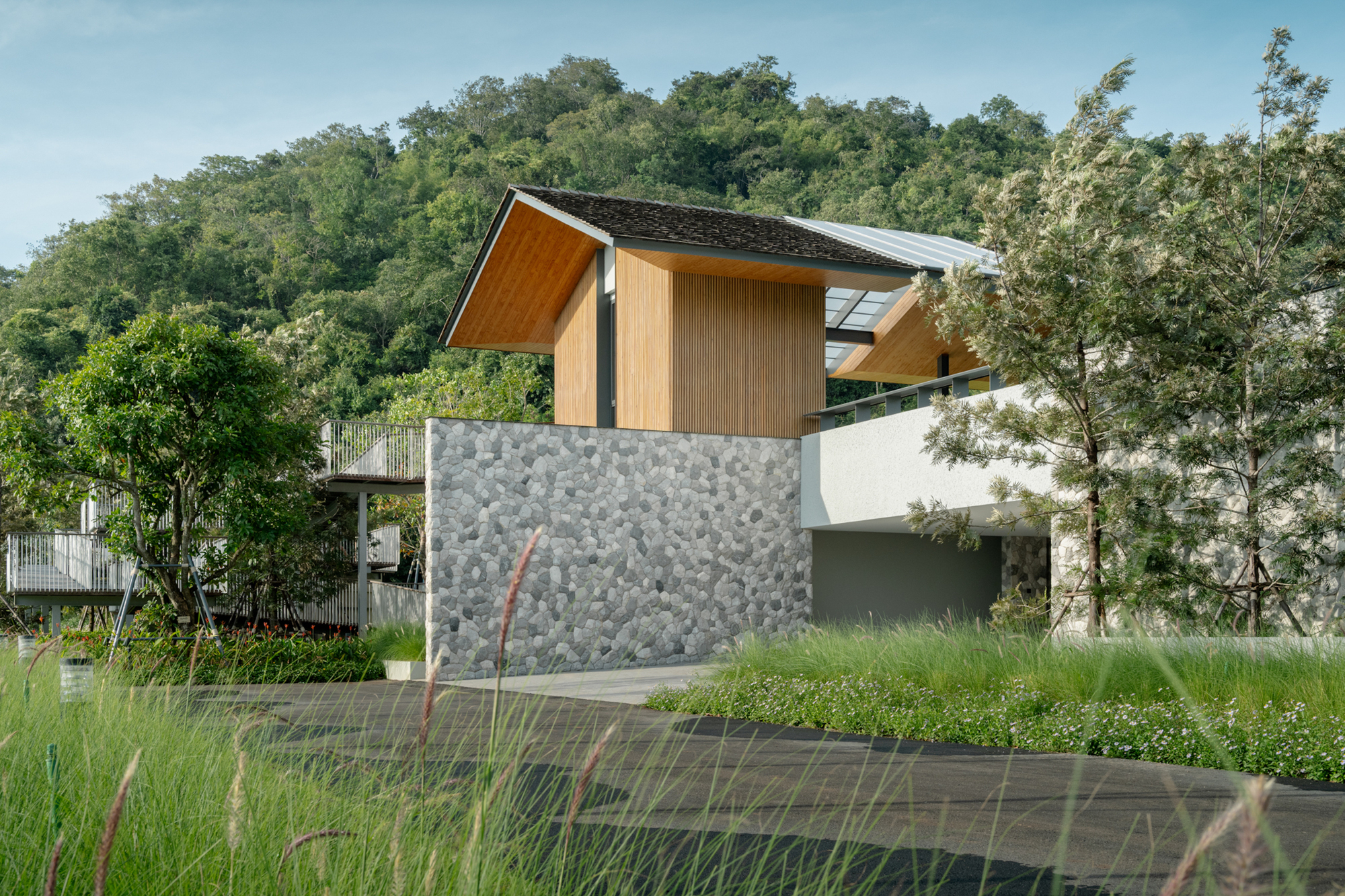 Timber-clad upper volume above a pebble-faced stone wall, with a forested hillside behind and grasses in the foreground