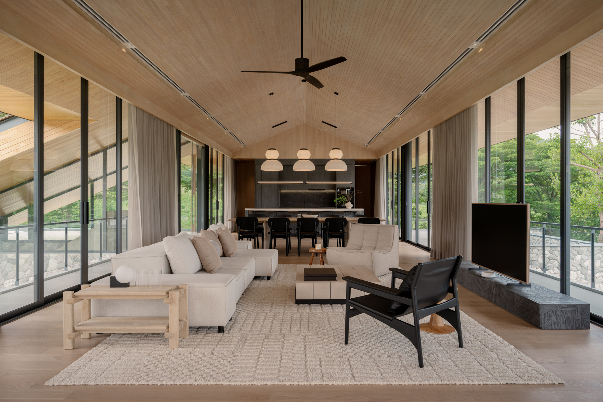 Living and dining hall under a pitched timber ceiling, with floor-to-ceiling glazing opening to green treetops outside