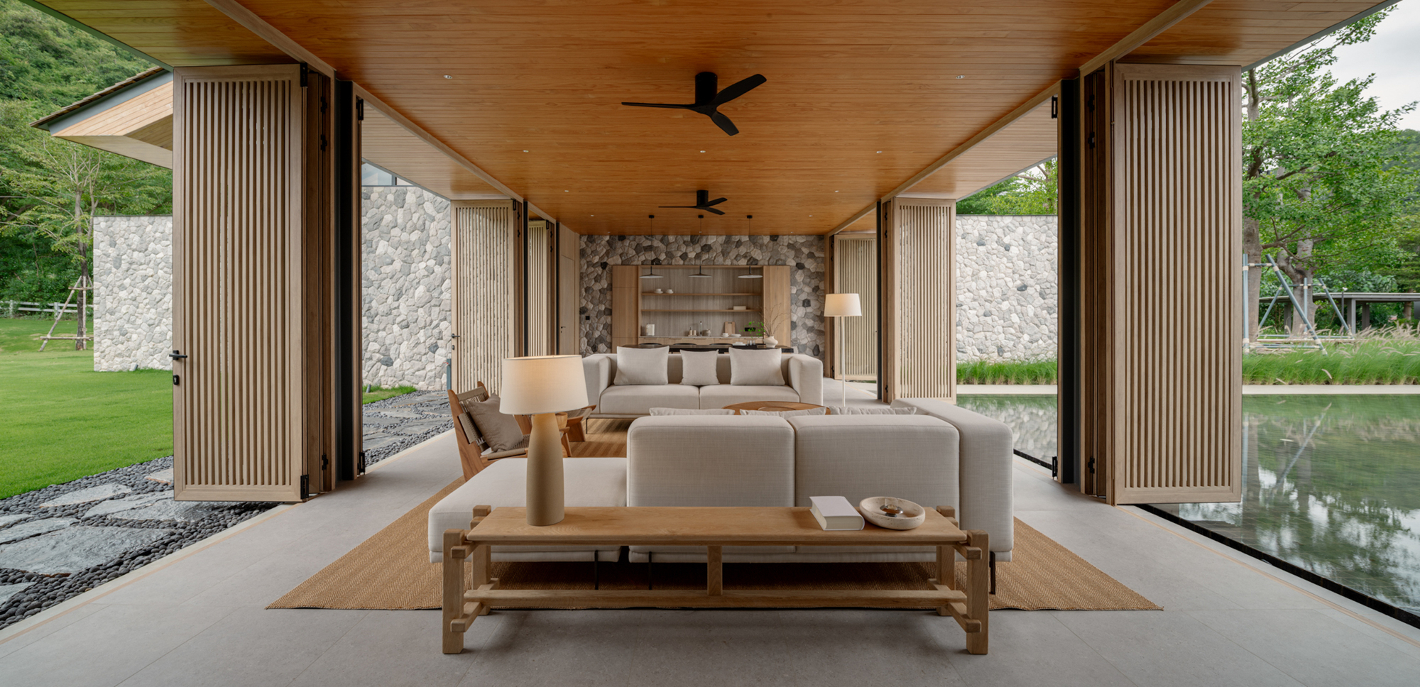 Open sitting pavilion with folded slatted timber shutters, stone walls and a reflecting pool seen at the right edge in soft overcast light