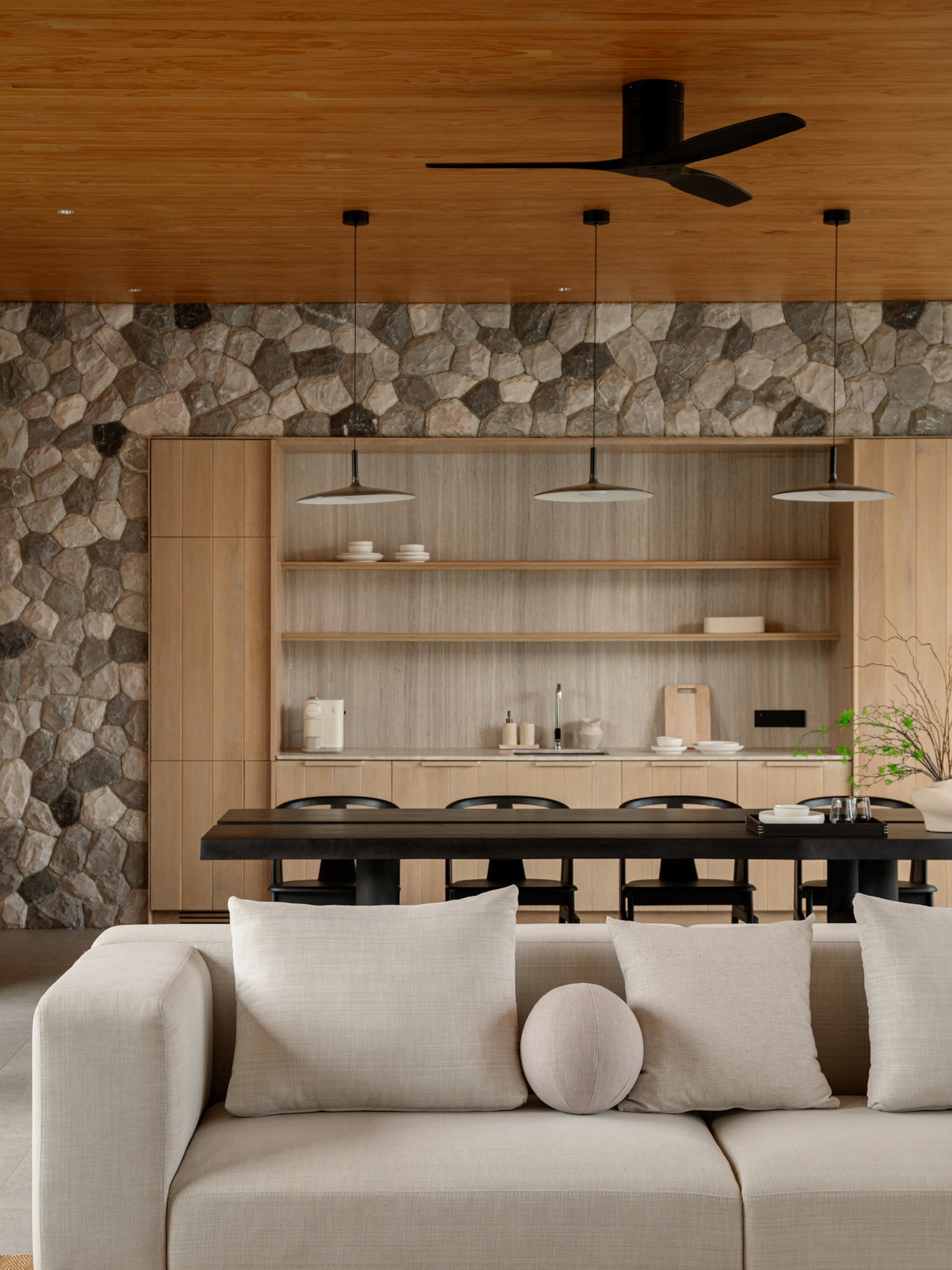 Kitchen wall of oak cabinetry and open shelves against a rubble stone backdrop, with pendant lamps over a black dining table