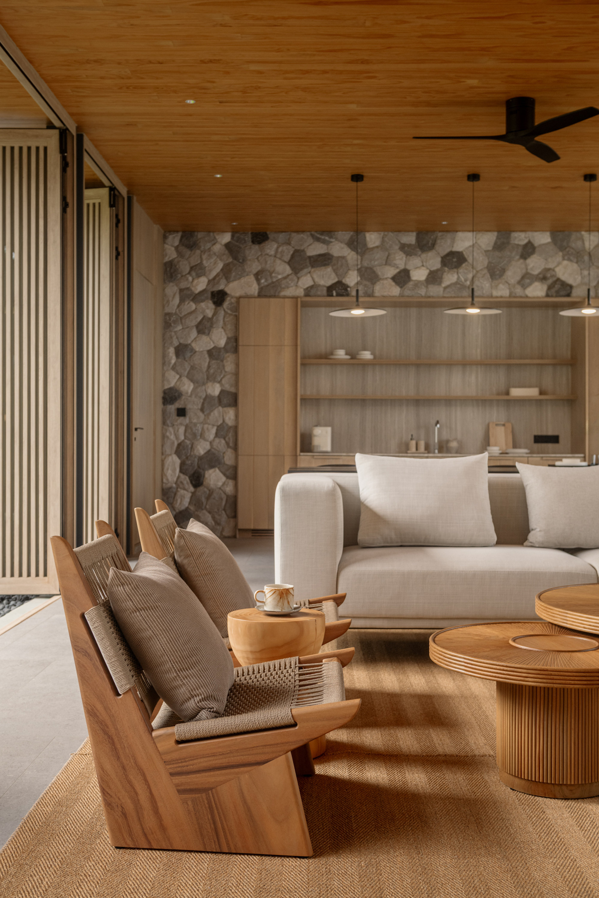 Living area with teak lounge chairs, a ribbed round table and a pale sofa before a stone feature wall and timber kitchen shelving