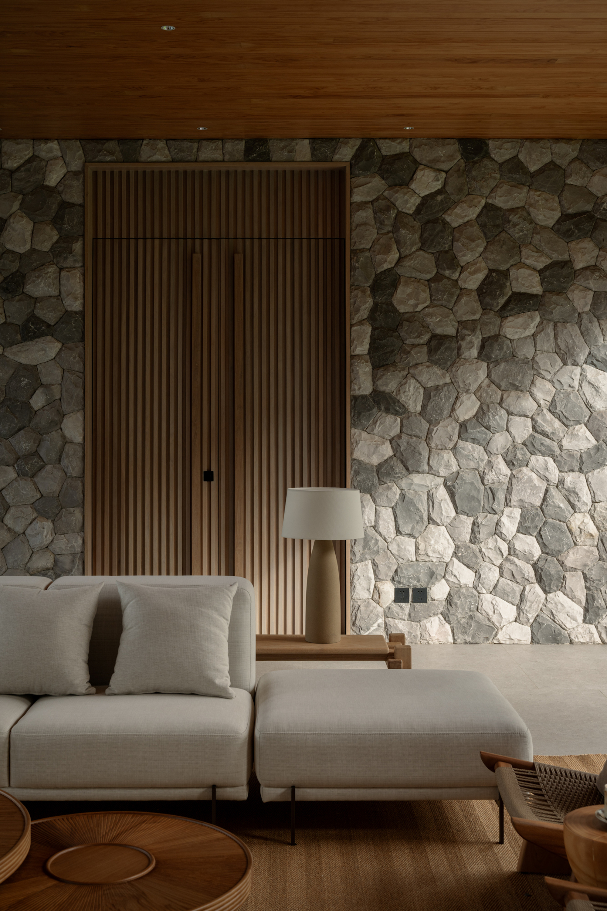 Slatted timber panel door set into a pebble-faced stone wall, with a lamp and cream sofa in dim evening light