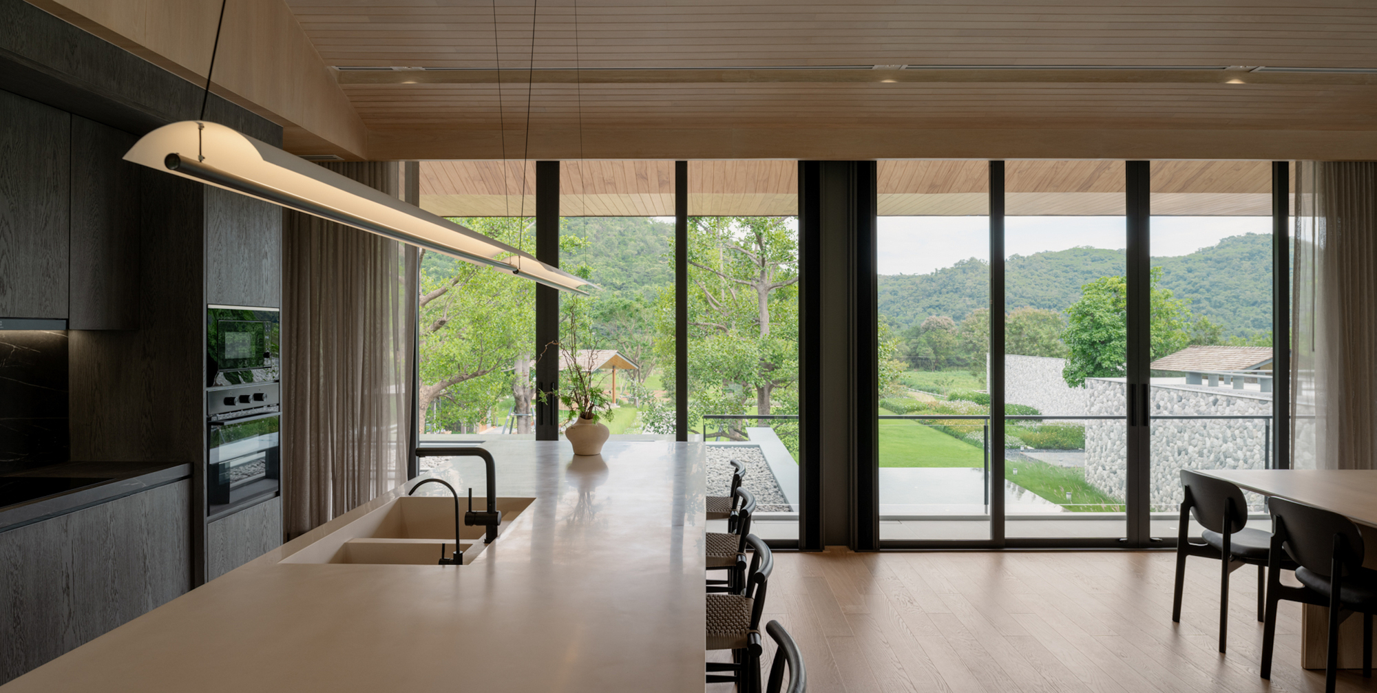 Kitchen island with a pendant light, looking through floor-to-ceiling glass to a green lawn and forested hills under overcast sky