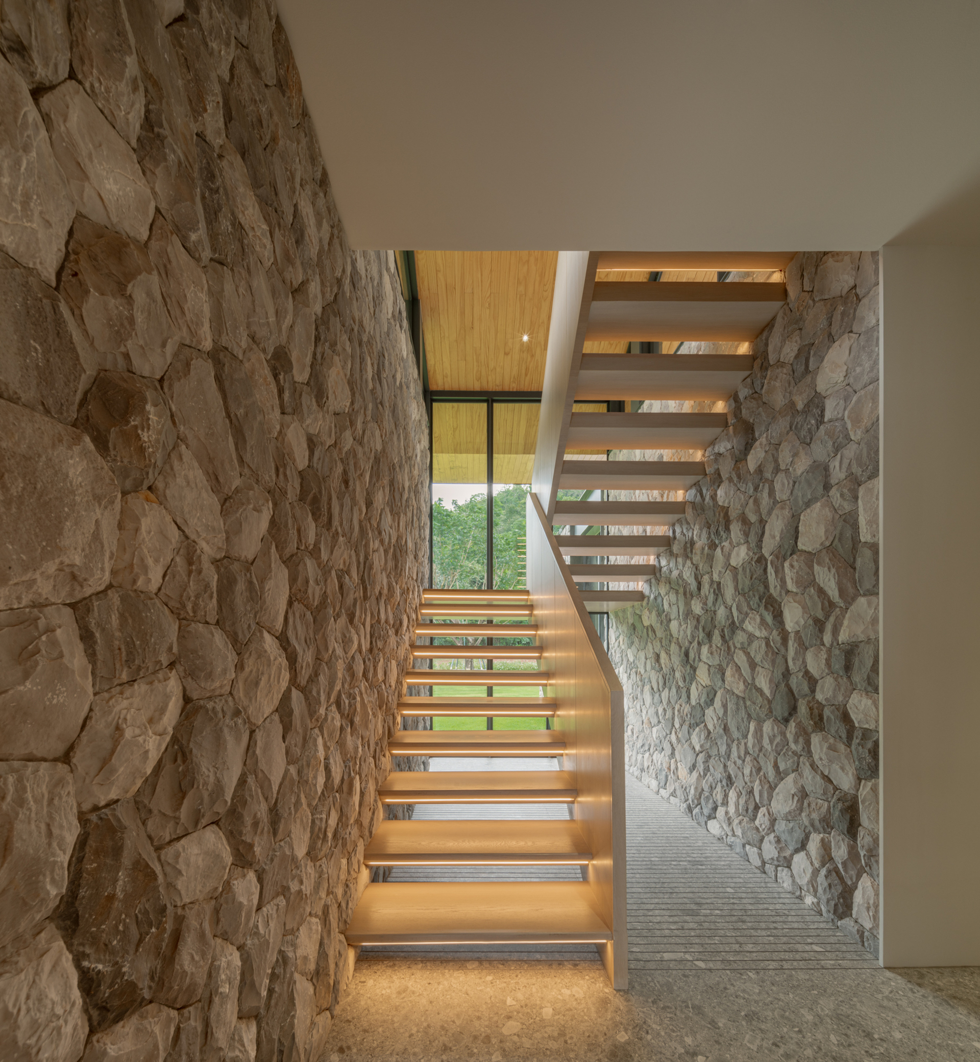 Floating timber stair with lit treads rising between two rough stone walls toward a glazed opening and trees