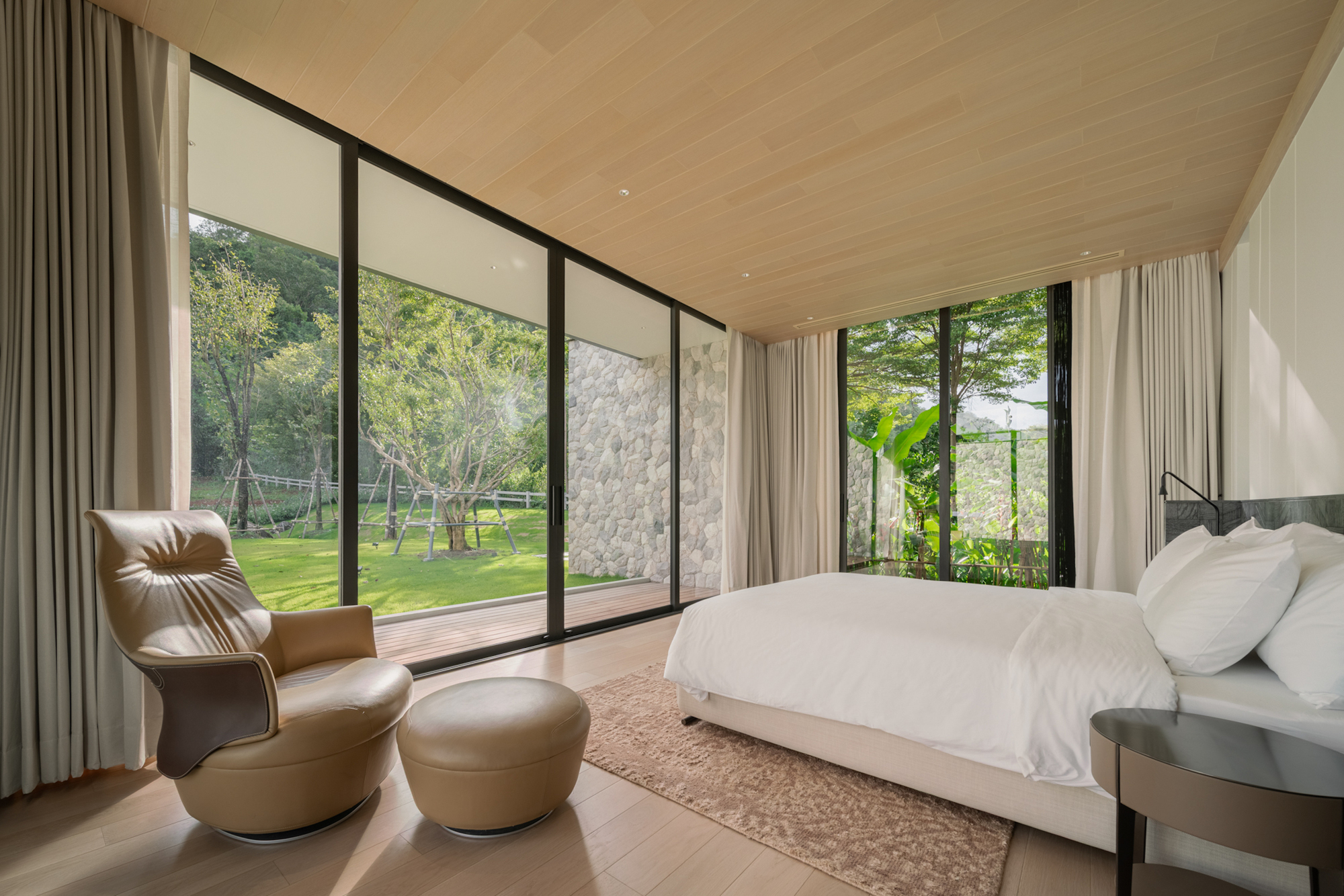 Bedroom with a leather armchair and ottoman, full-height sliding glass and a timber deck opening onto a sunlit lawn