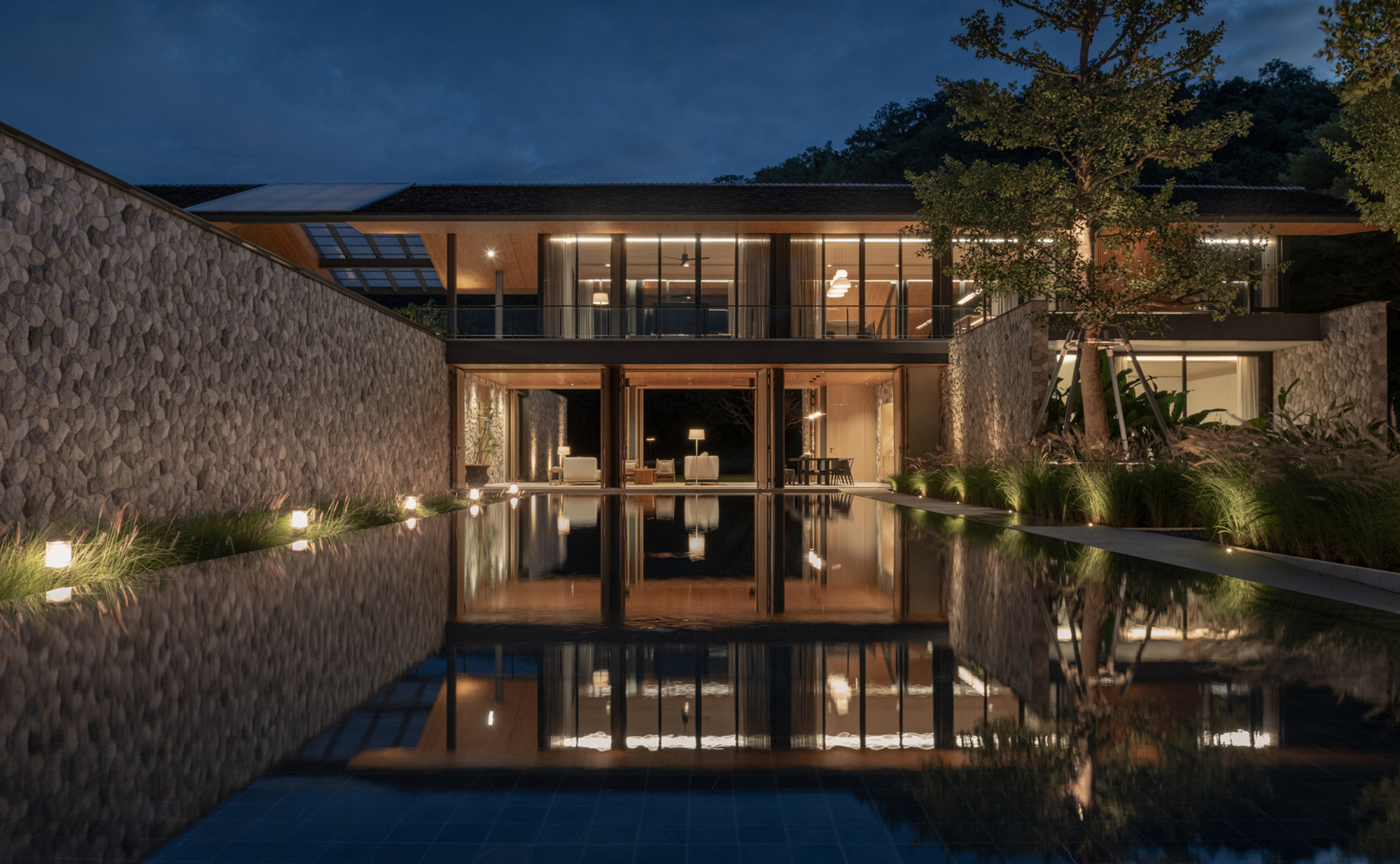 Stone wall and glazed two-storey pavilion reflected in a long still pool at dusk, grass lights glowing