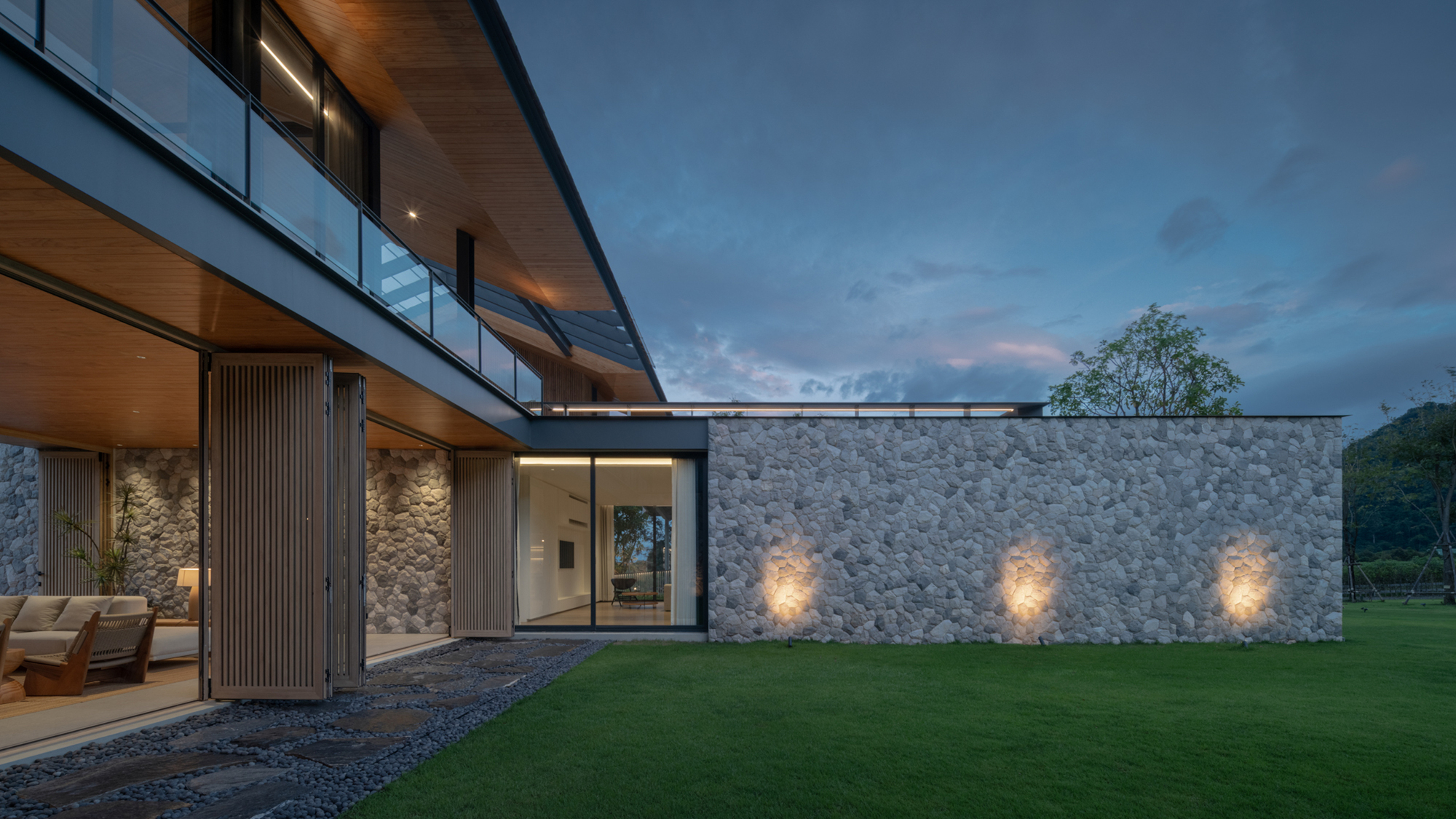 Stone-clad garden wall lit by three uplights beside a timber-lined balcony and sliding screens at twilight