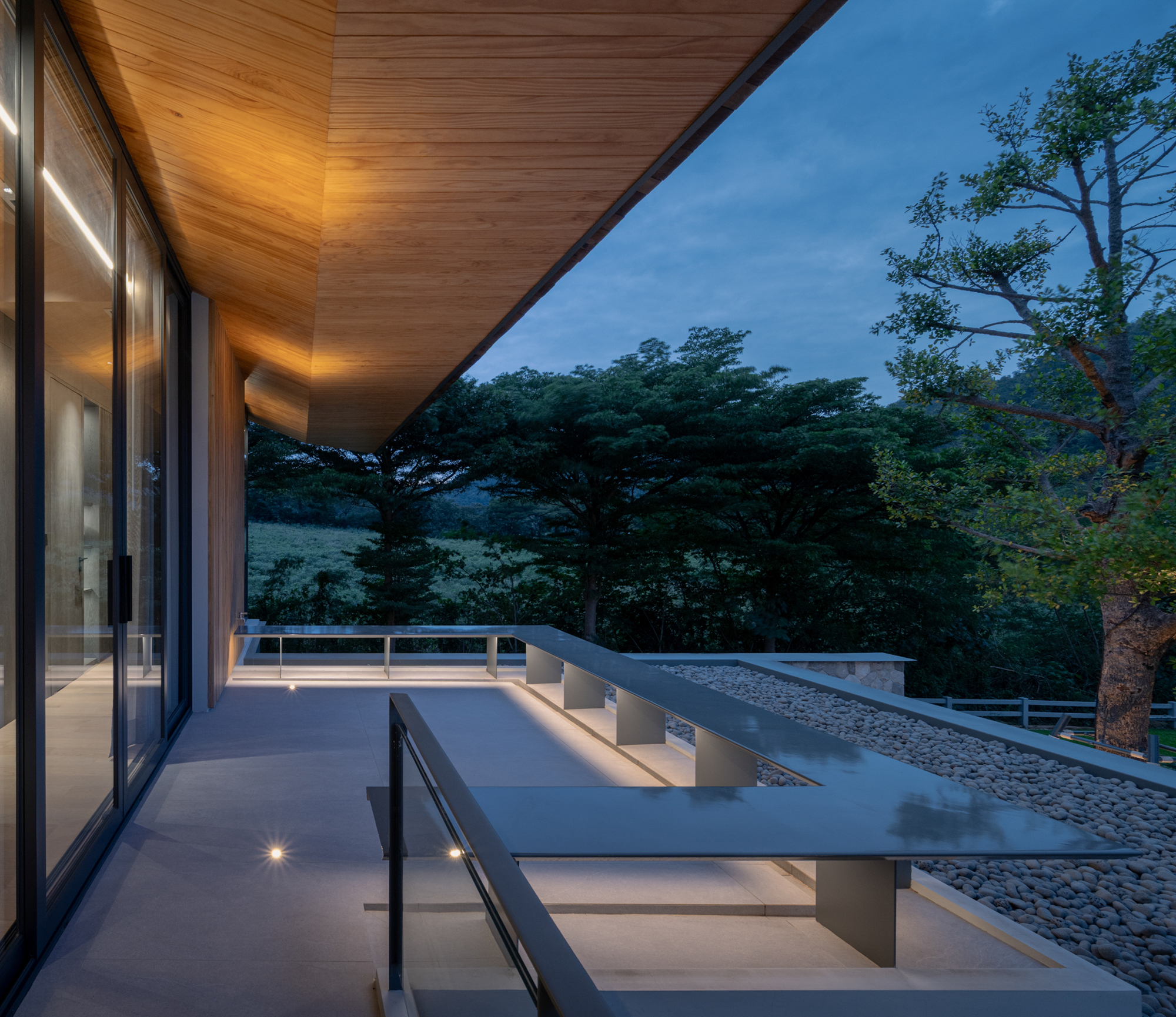 Terrace under a timber soffit with a steel bench, glass balustrade and pebble roof edge beside a large tree at dusk