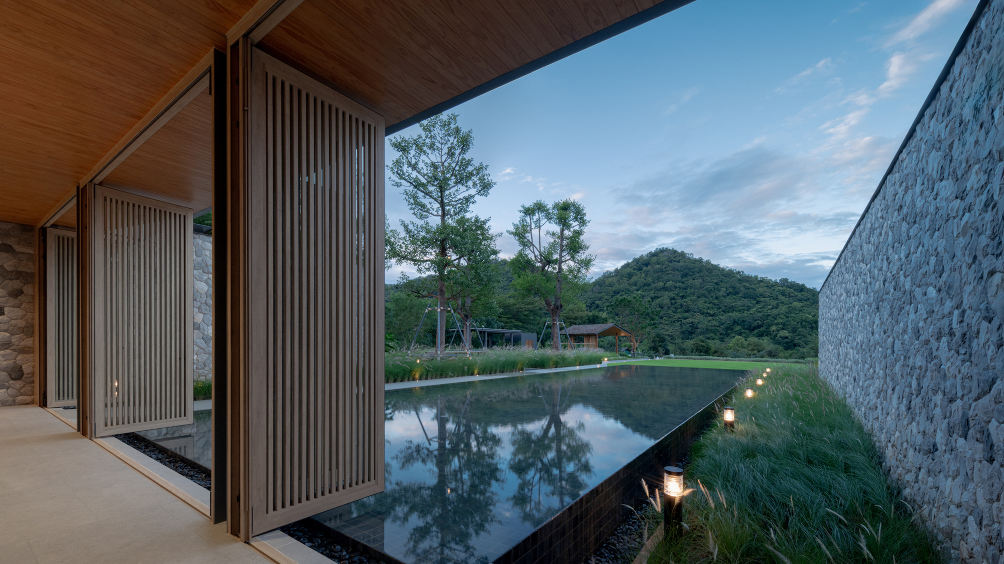 Folding slatted timber screens open onto a reflecting pool, stone wall and grass bed with a forested hill beyond