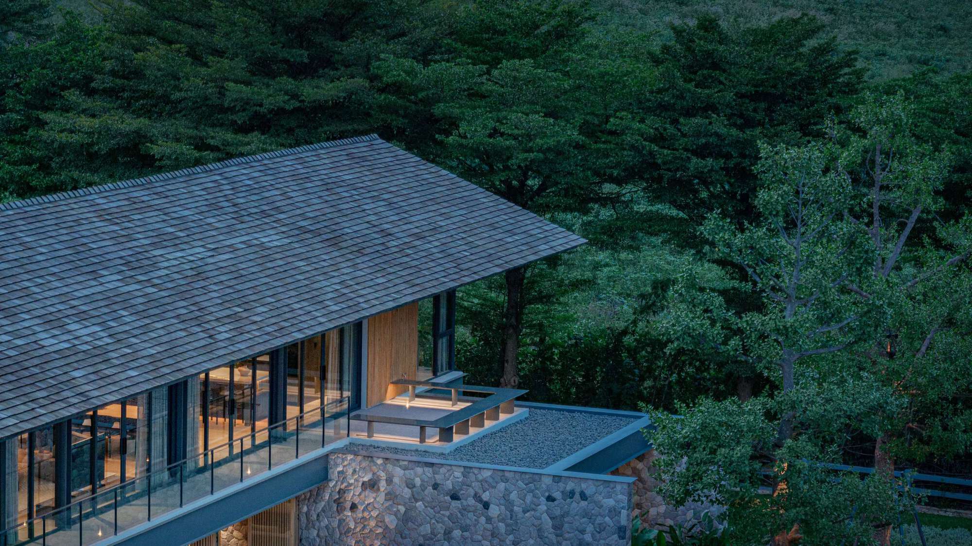 Aerial view of the shingled pitched roof, glazed upper floor and stone base with a rooftop deck, among trees at dusk
