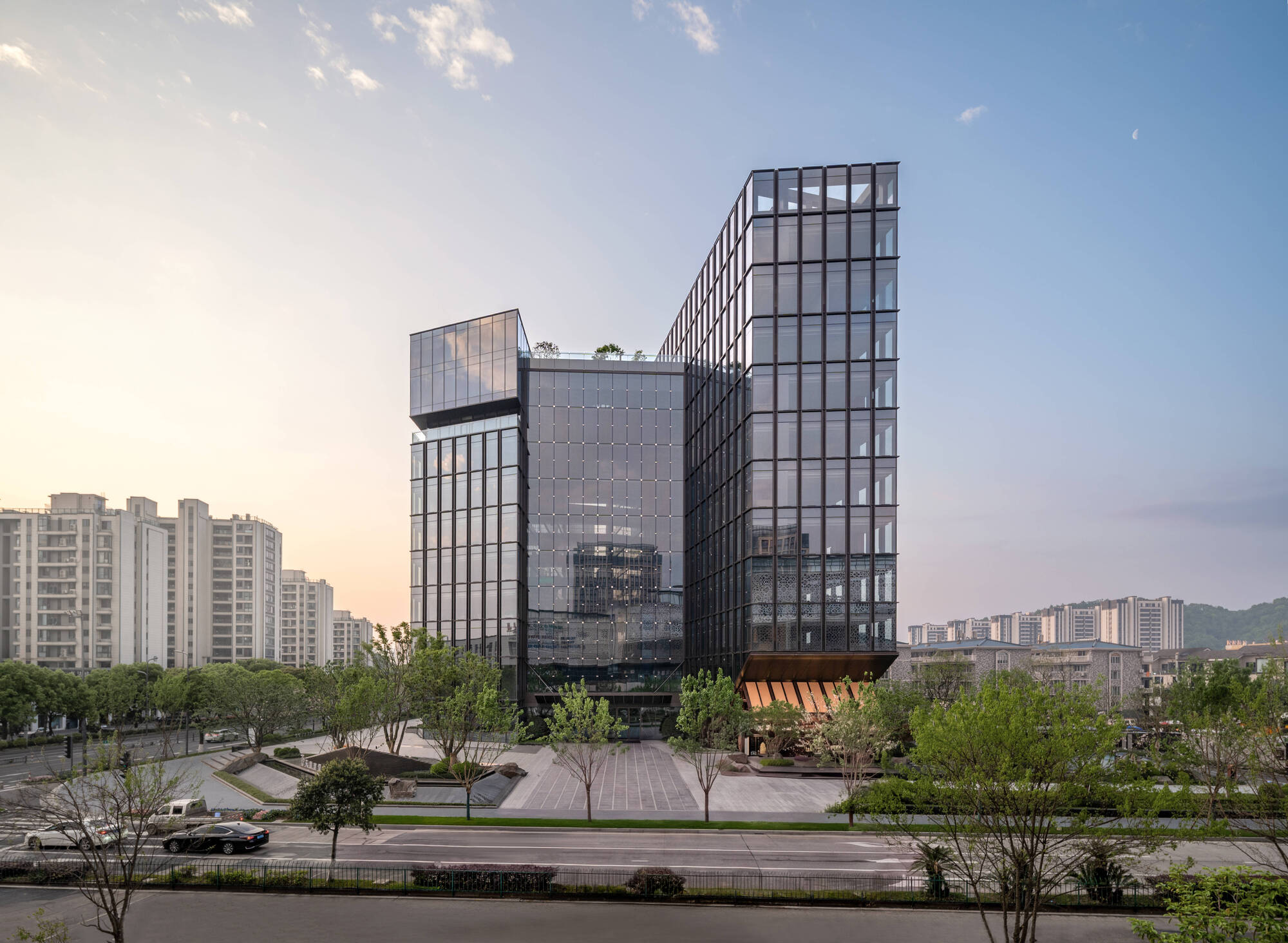 Two glass office towers with a bronze-lit canopy beyond a paved plaza and young trees at dusk
