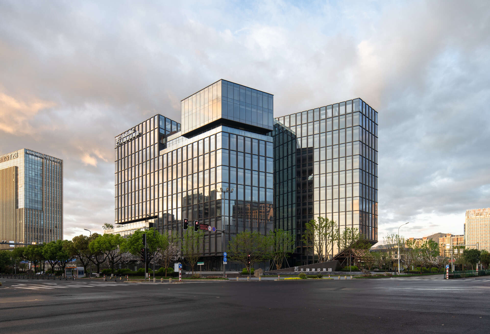 Corner view of stepped glass towers from an empty intersection under a cloudy sky lit by low sun