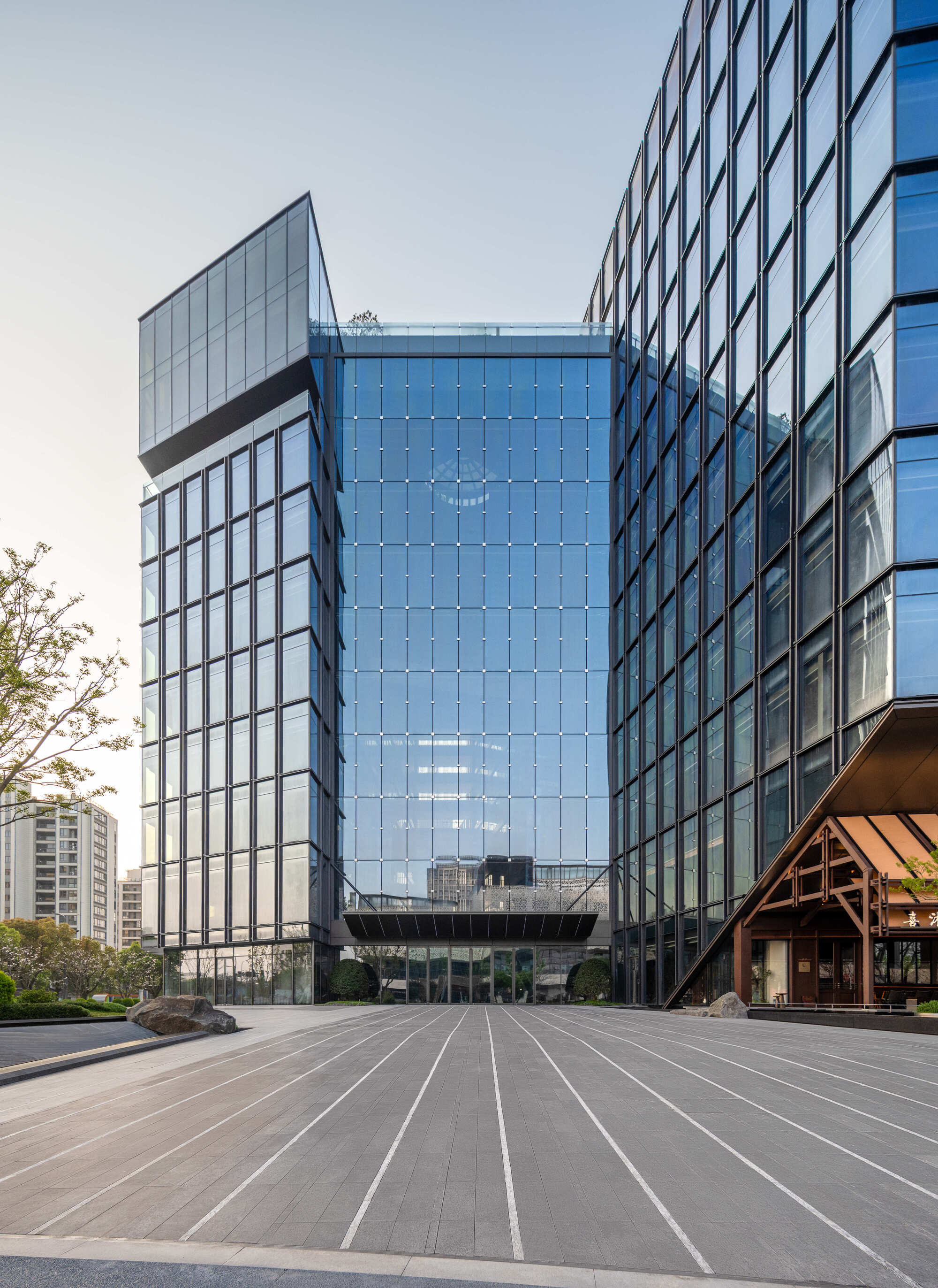 Glass tower facades framing a wide paved plaza with linear stripes, with a timber-framed entrance canopy at right