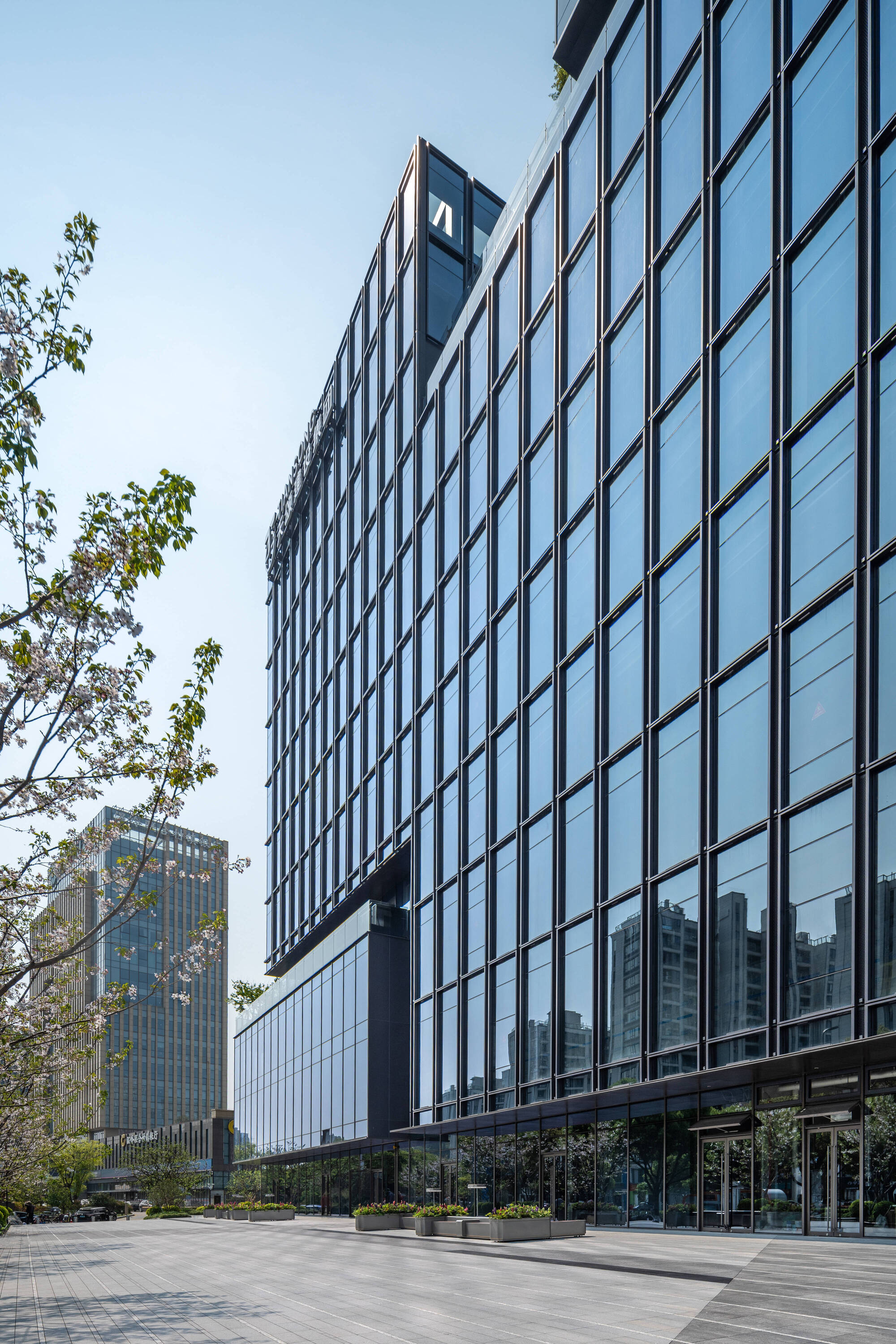 Low-angle view along the grid-patterned glass curtain wall above a paved plaza with planters and blossoming branches