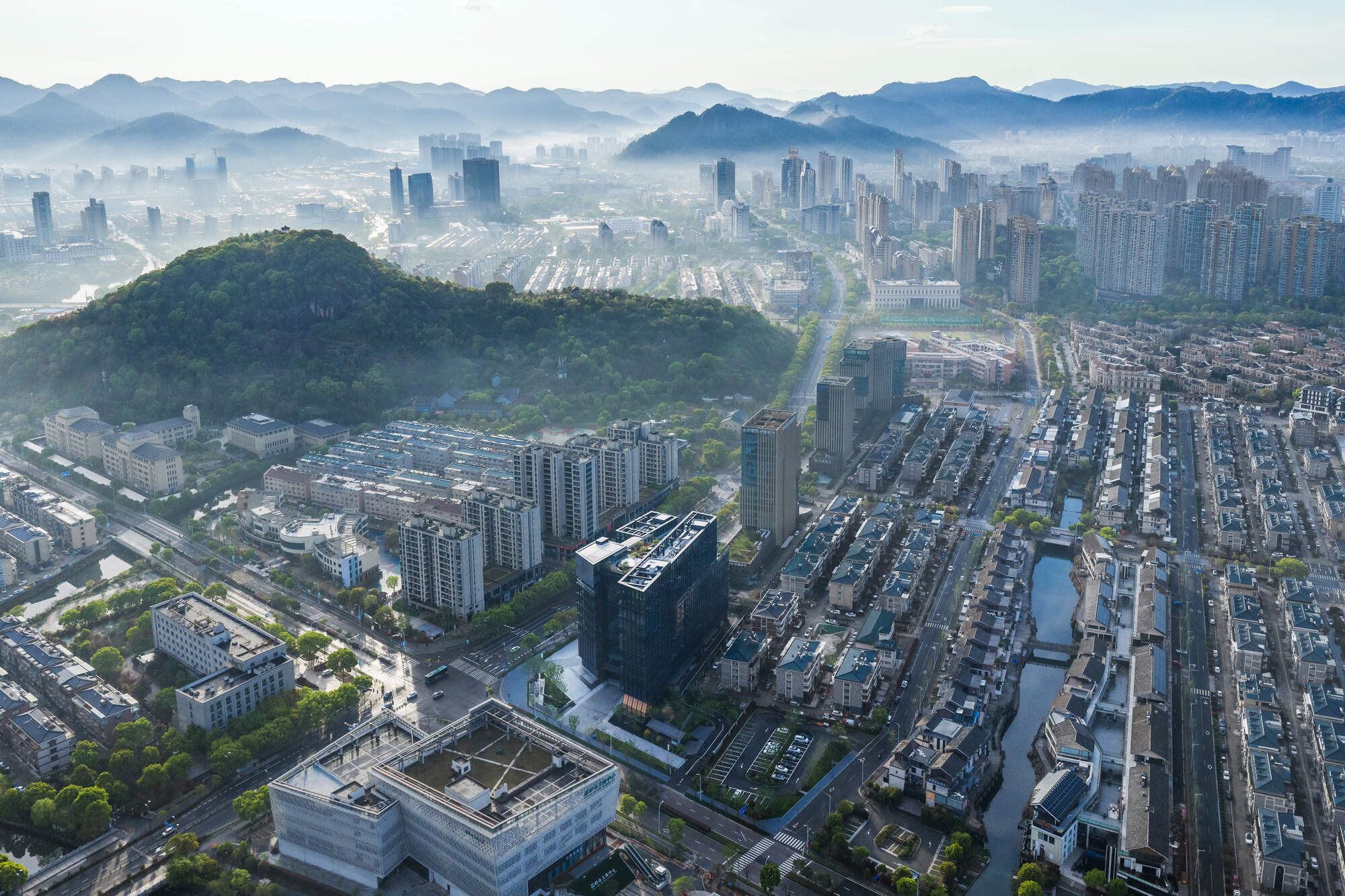 Aerial view of the dark glass tower amid canal-side housing, with a wooded hill and misty mountains behind at dawn