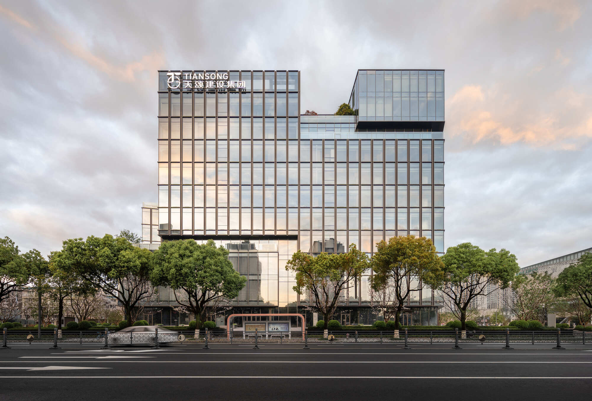Street-level view of the glass curtain wall facade with a stepped upper volume, framed by trees under a pink-tinged cloudy sky