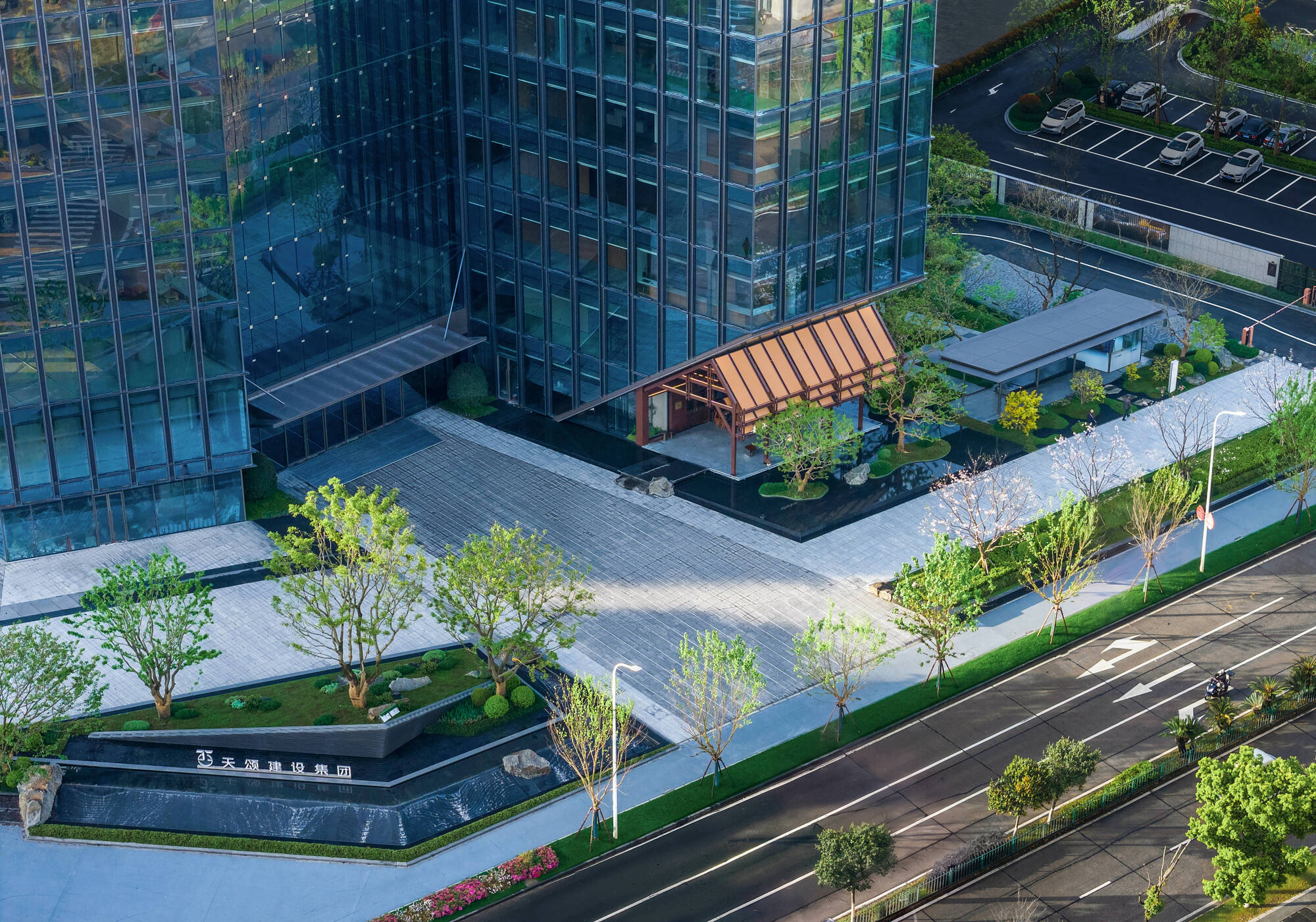 Aerial view of the paved plaza with a timber-framed pavilion, reflecting pool and young trees beside the glass tower