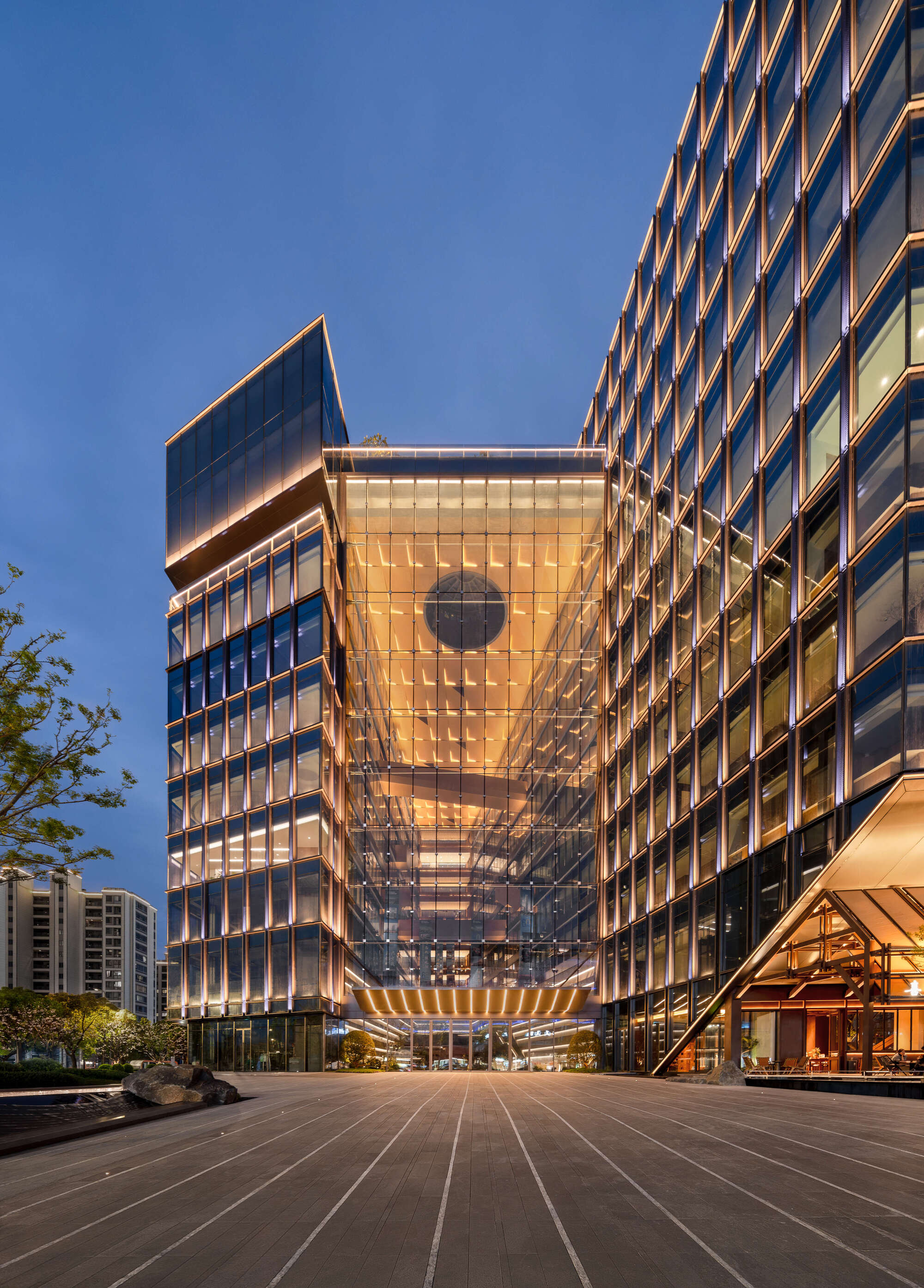 Glass atrium void between two stepped towers, lit amber at dusk above a timber-decked plaza with a circular opening overhead
