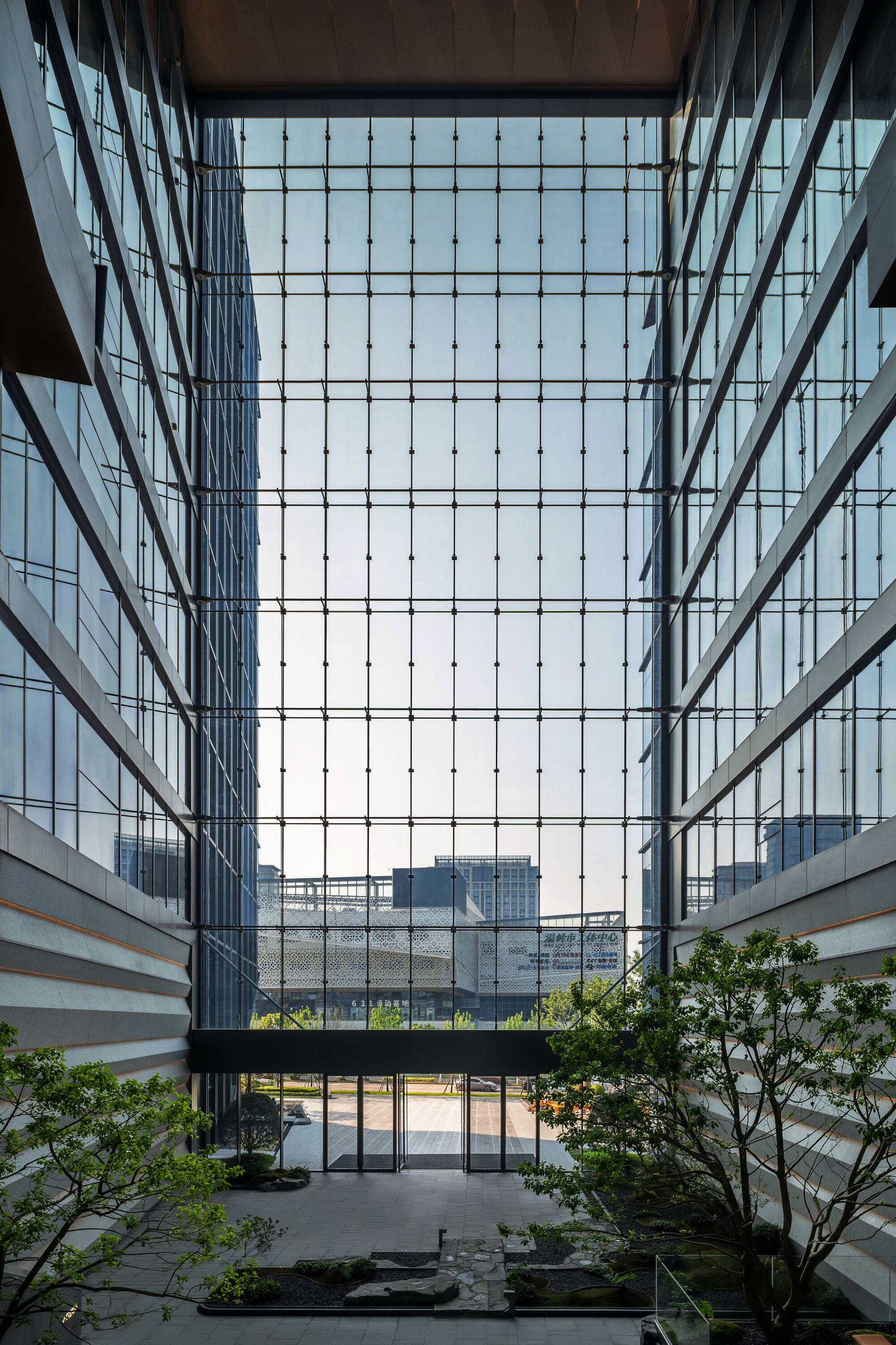 Tall glazed atrium wall framed by two tower facades, with a planted stone garden and trees at its base