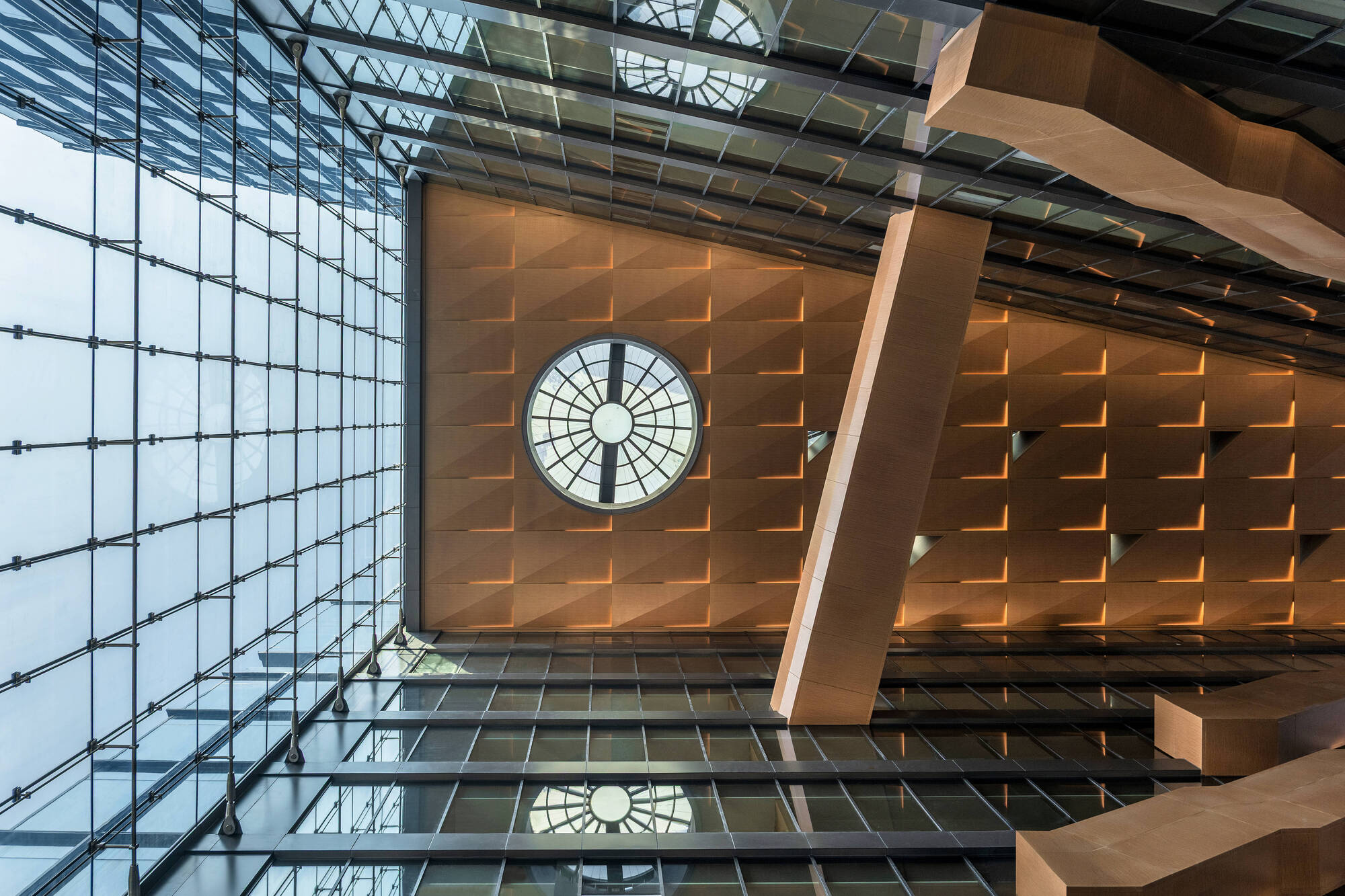 Upward view of the atrium's timber-clad wall, round skylight window and angled column beneath a glass roof with warm lighting