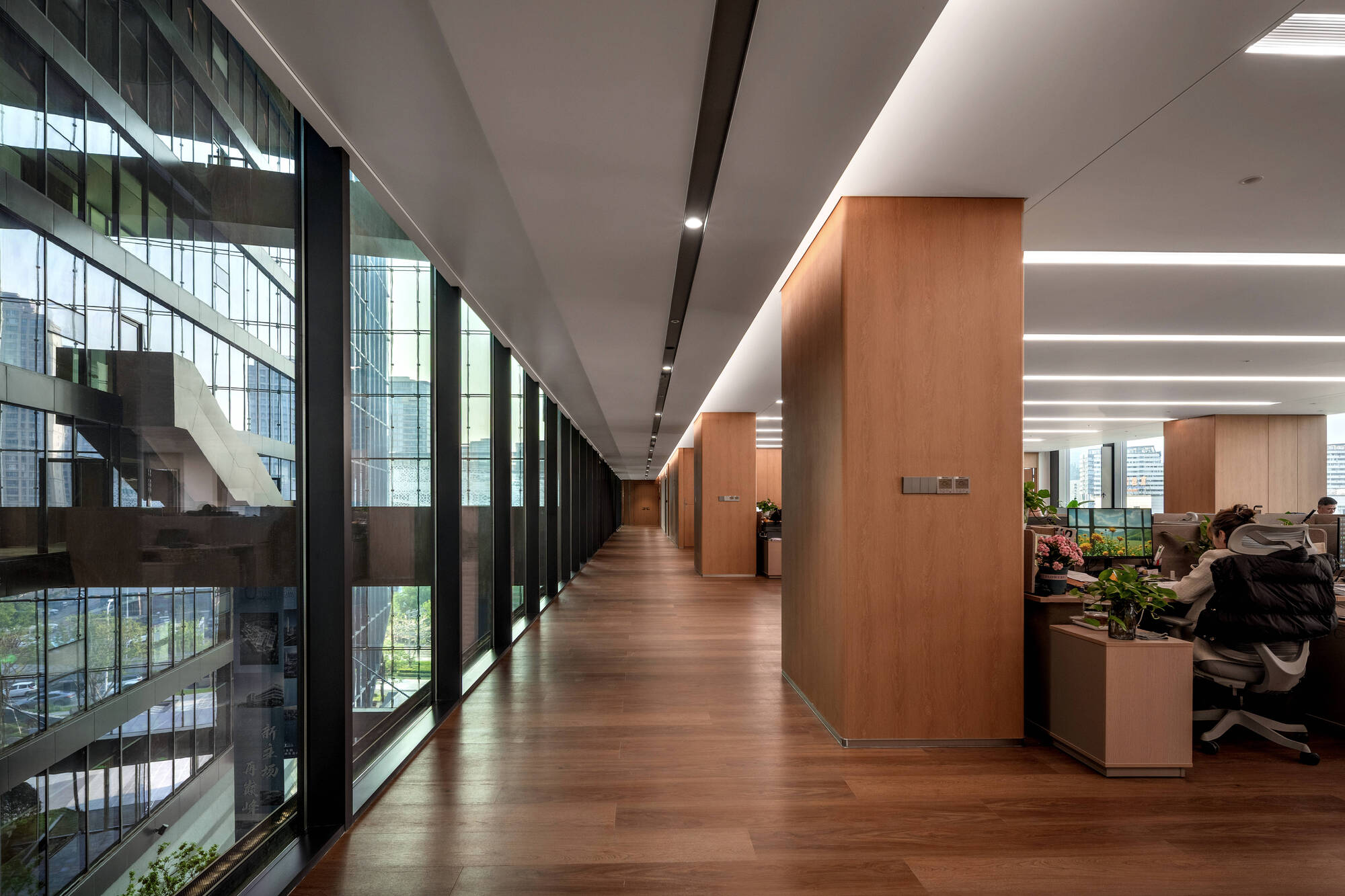 Office floor corridor with timber-clad columns and wood flooring running beside a glass wall, a worker seated at a desk