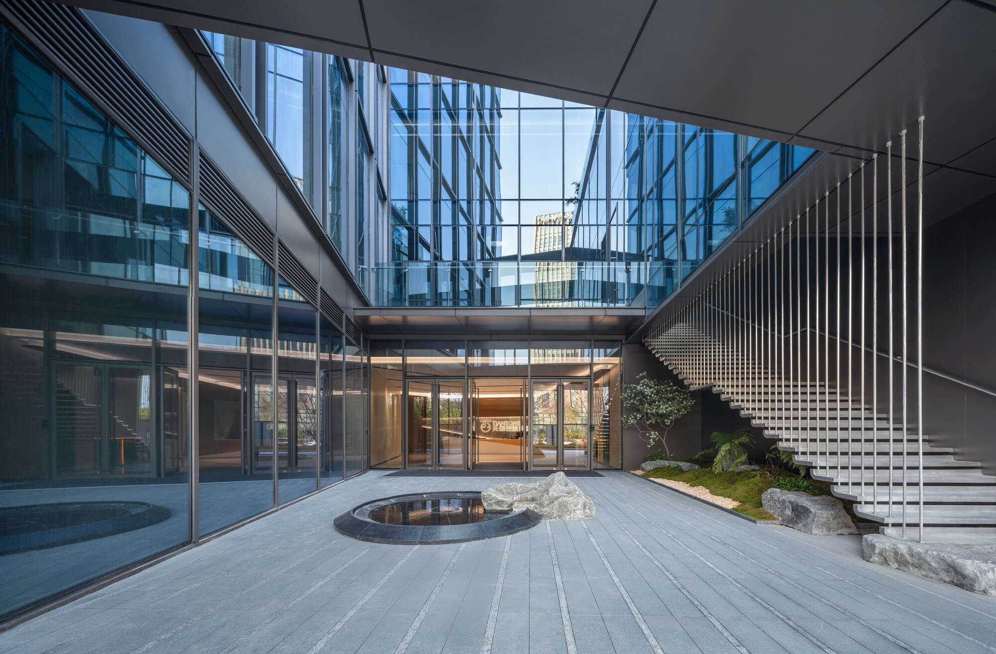 Paved entrance courtyard with a round pool, boulders, planted bed and a floating stair hung with steel rods in daylight