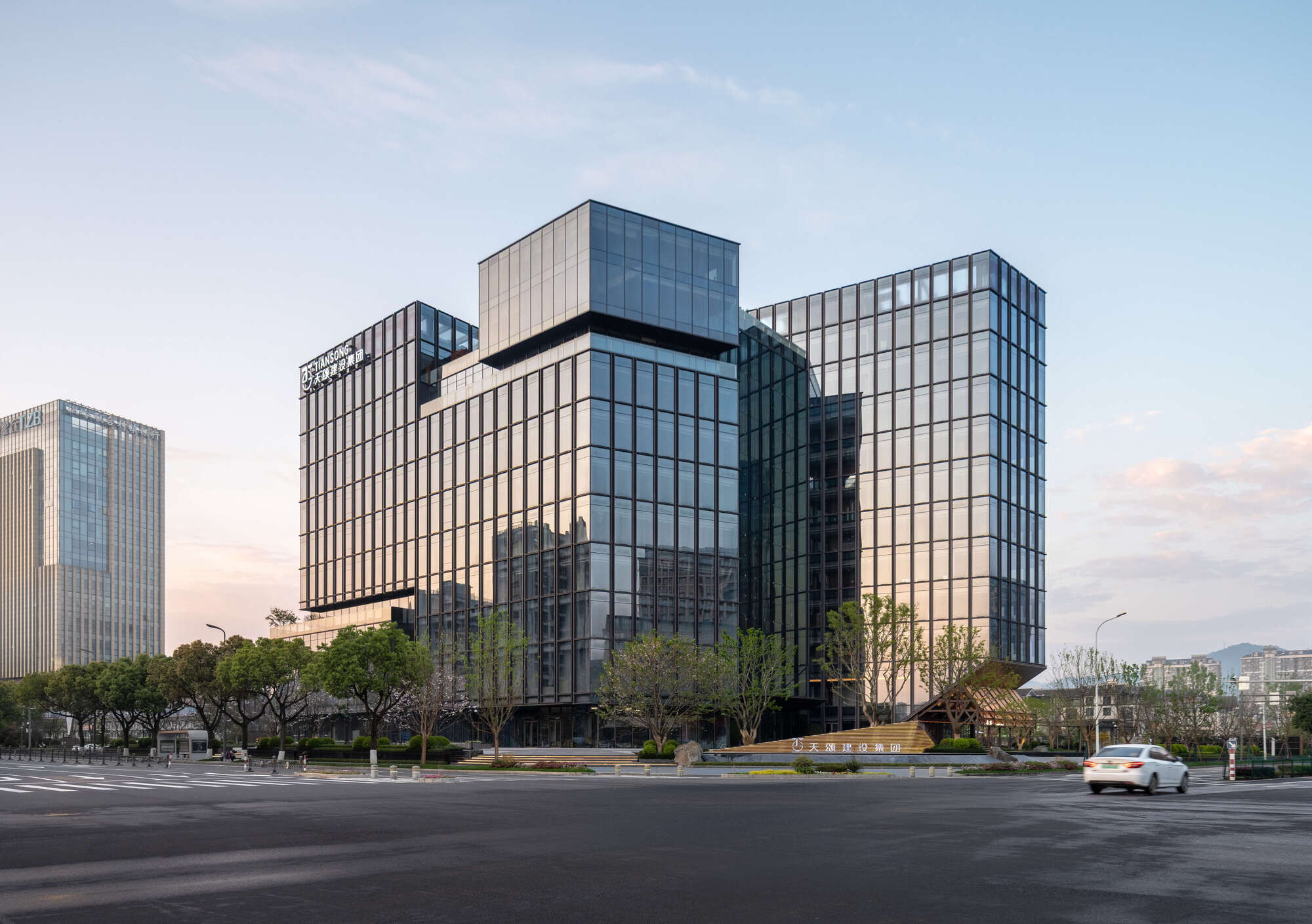 Street view of the stepped glass office volumes at dusk, with a white car passing and trees along the plaza