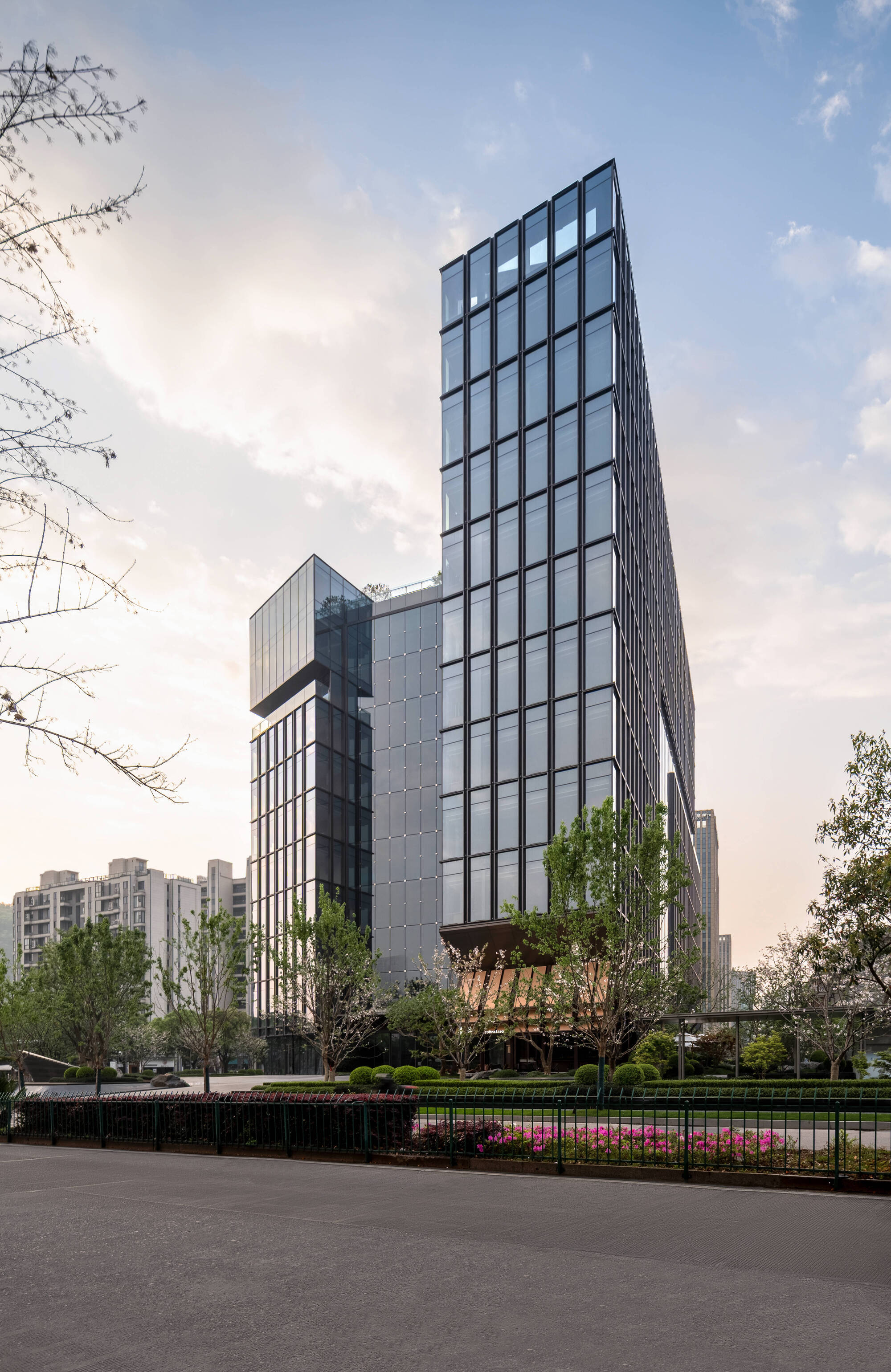 Tall glass tower with a stepped lower volume rising behind a flowering planted bed and bare branches at dusk