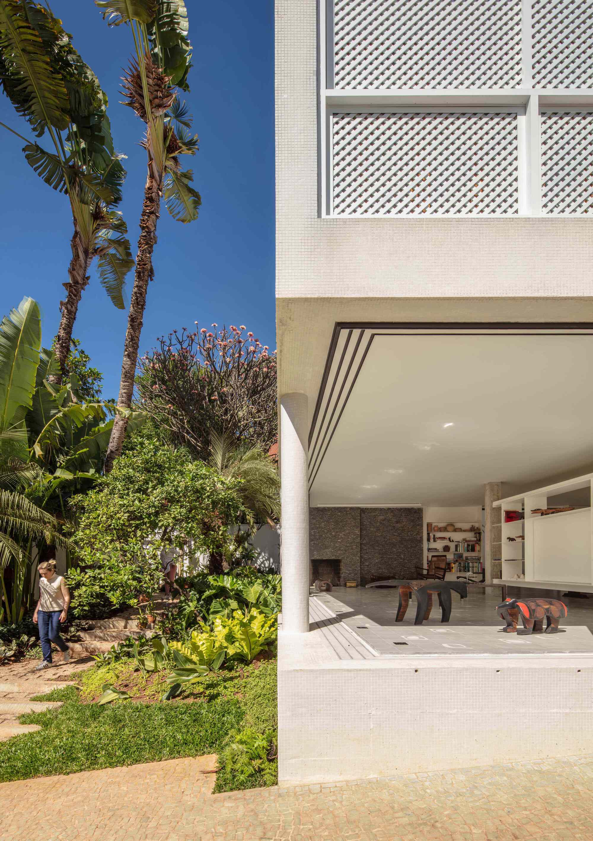 Lattice-screened upper floor over an open living room, with a woman walking a garden path beneath tall palms