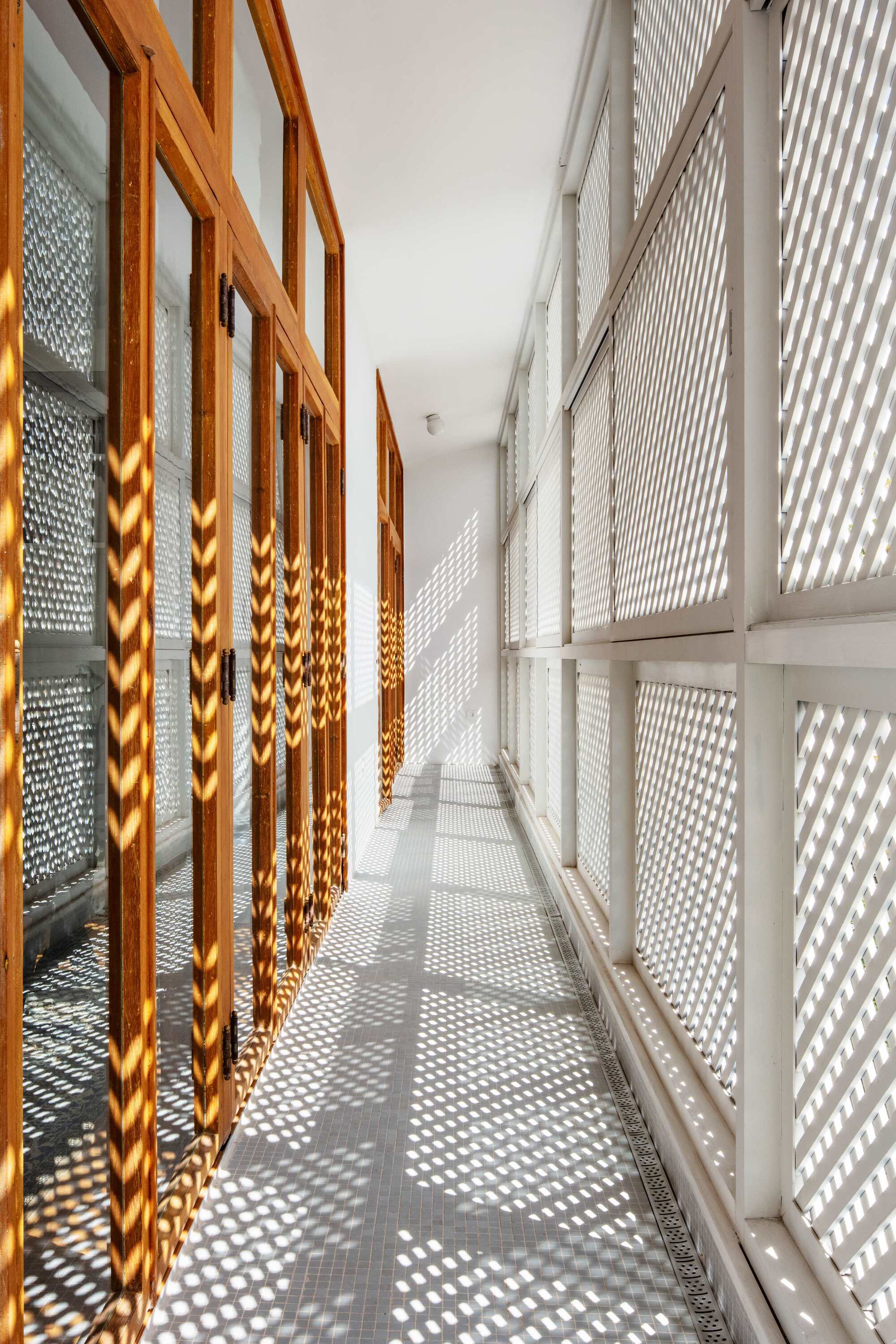 Narrow balcony corridor between timber doors and white lattice screens, with sunlight casting dotted shadows across the tiled floor