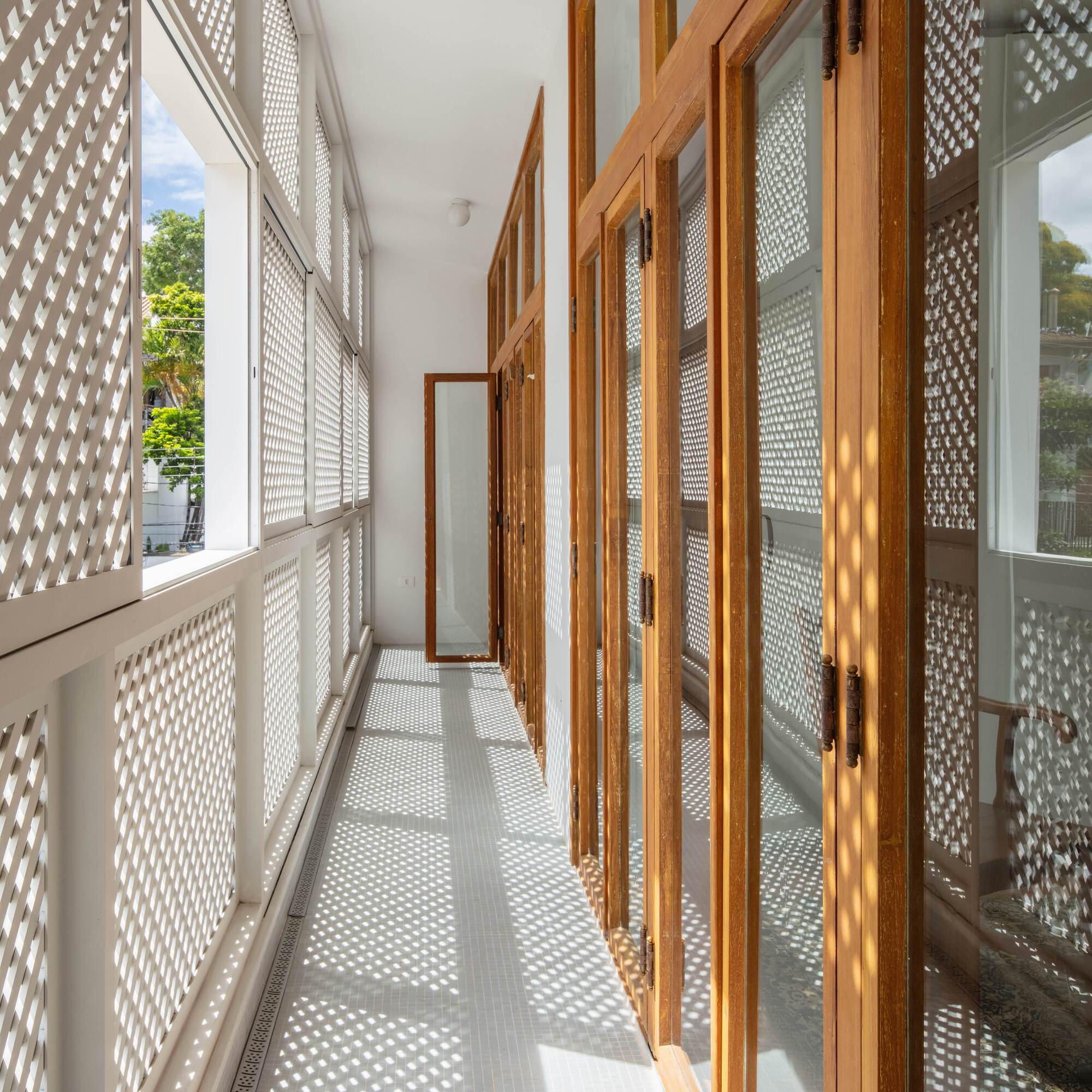 Tiled gallery running between white lattice panels and timber-framed glass doors, with patterned light falling along the floor