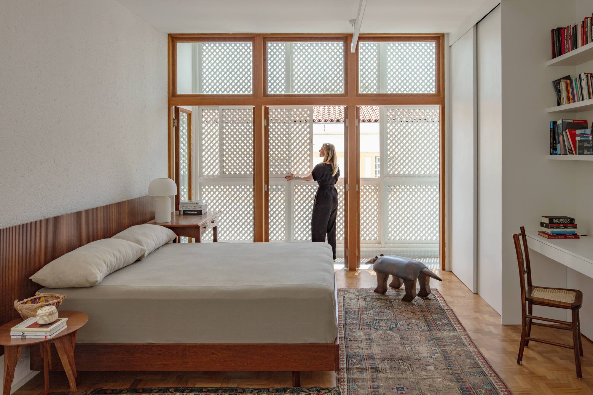 Woman standing at the open lattice doors of the bedroom, a carved wooden animal stool on the rug beside her