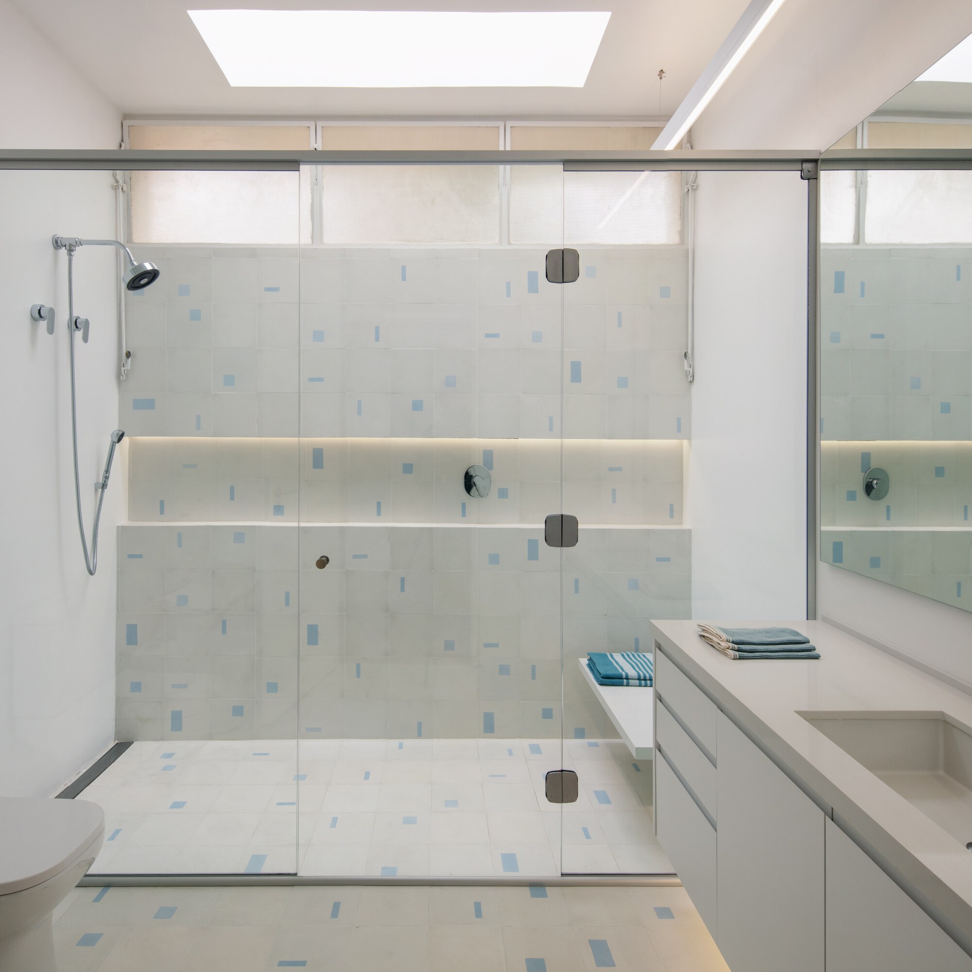 Walk-in glass shower with pale tiles speckled in blue, a lit niche and a skylight overhead