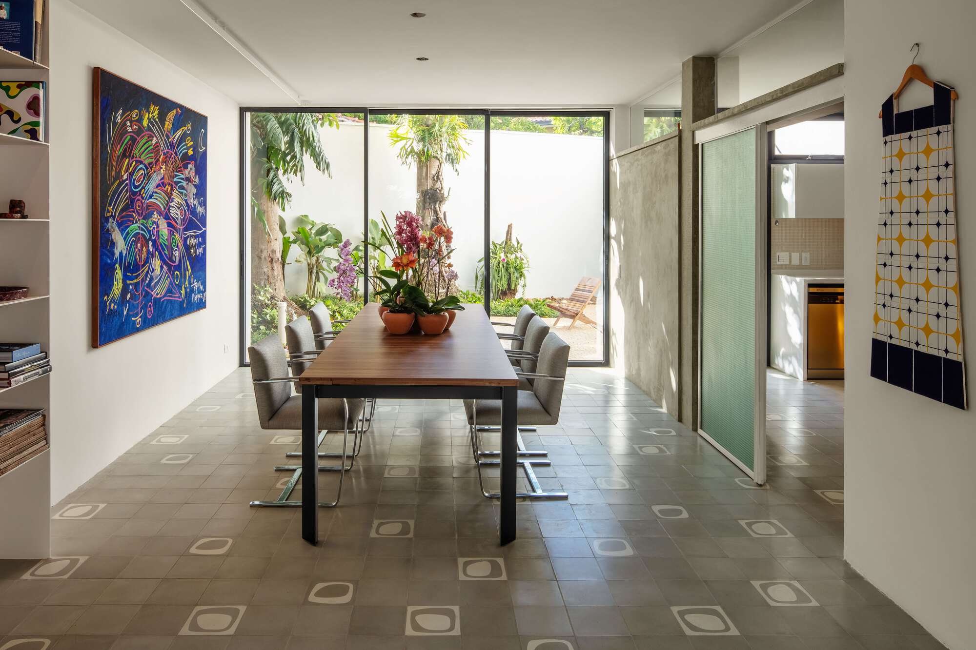 Dining table with orchids and steel-framed chairs facing a glass wall onto a palm garden in bright daylight