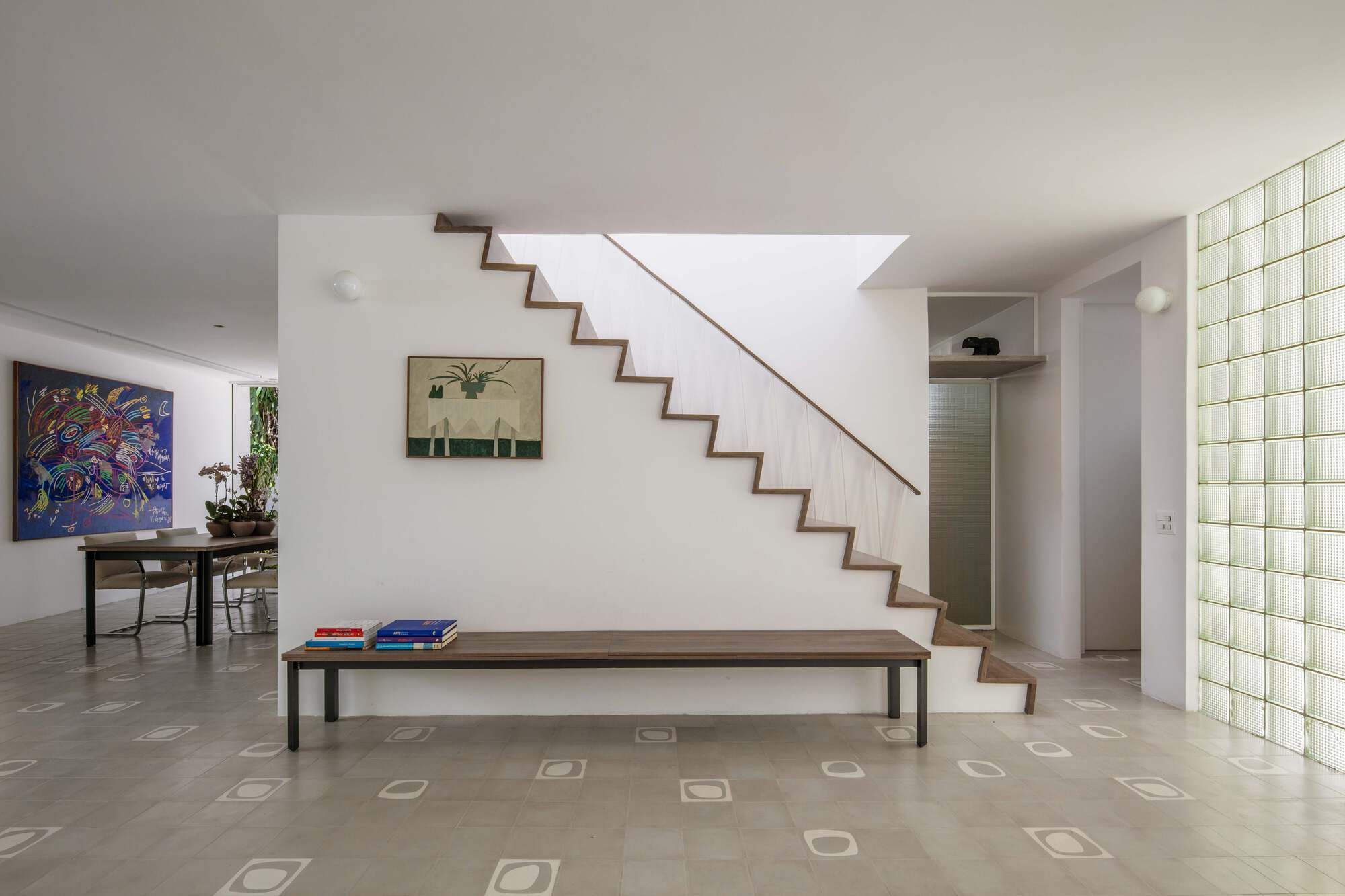Zigzag timber stair rising above a long wooden bench, with a glass block wall at right and skylight glow overhead