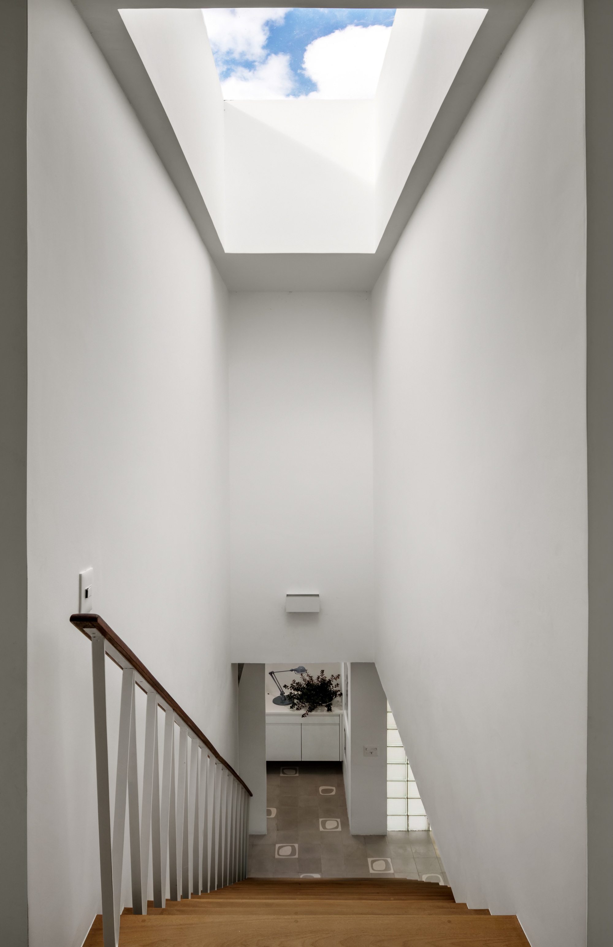 Stairwell looking down from a rectangular skylight framing blue sky and clouds, with white walls and a timber handrail