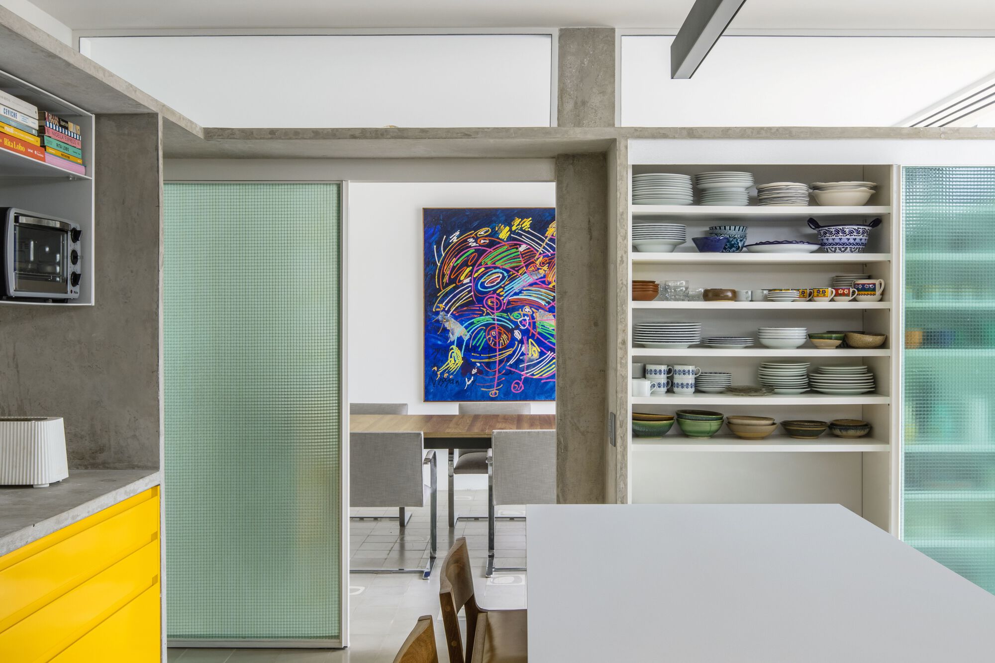 Open shelving of bowls and plates beside a green glass sliding door, framing a colorful painting in the dining room beyond