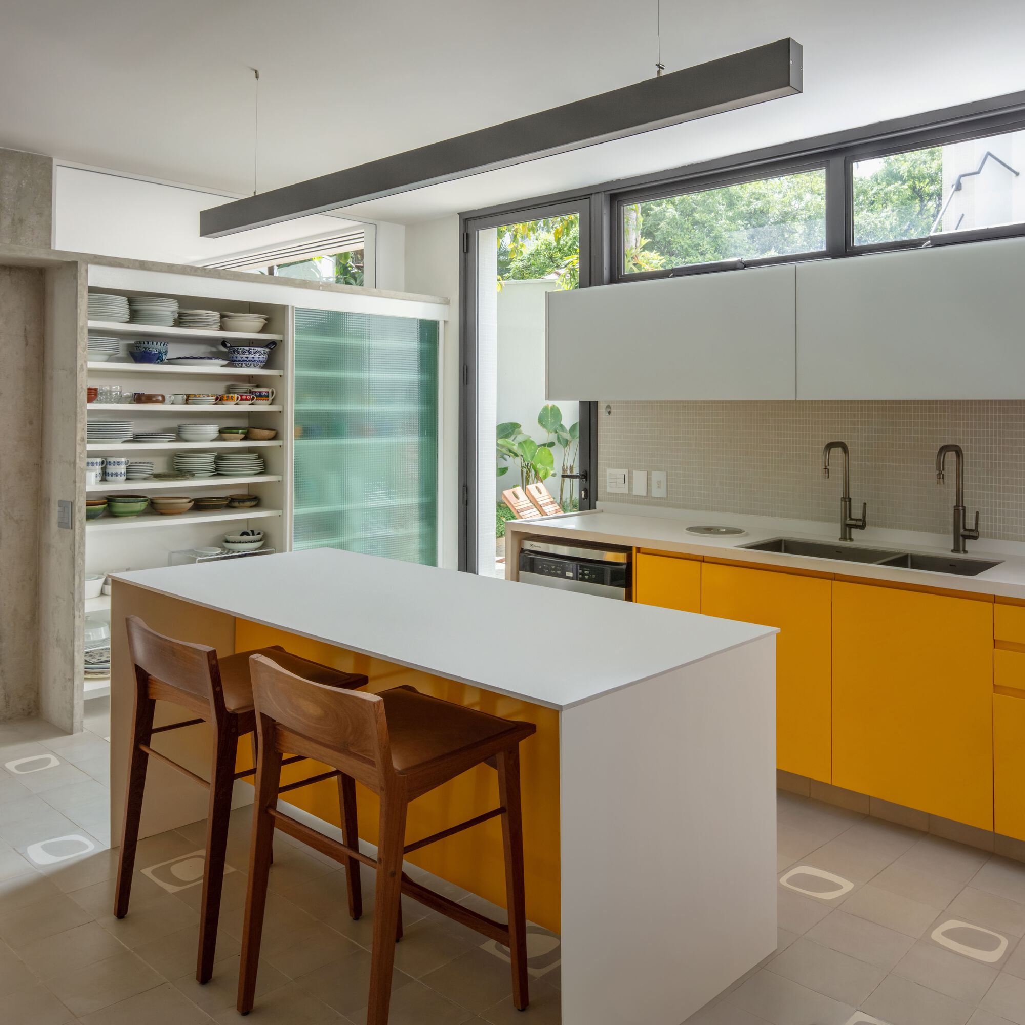 White kitchen island with two timber stools facing yellow cabinets and a double sink under high windows, with garden greenery outside