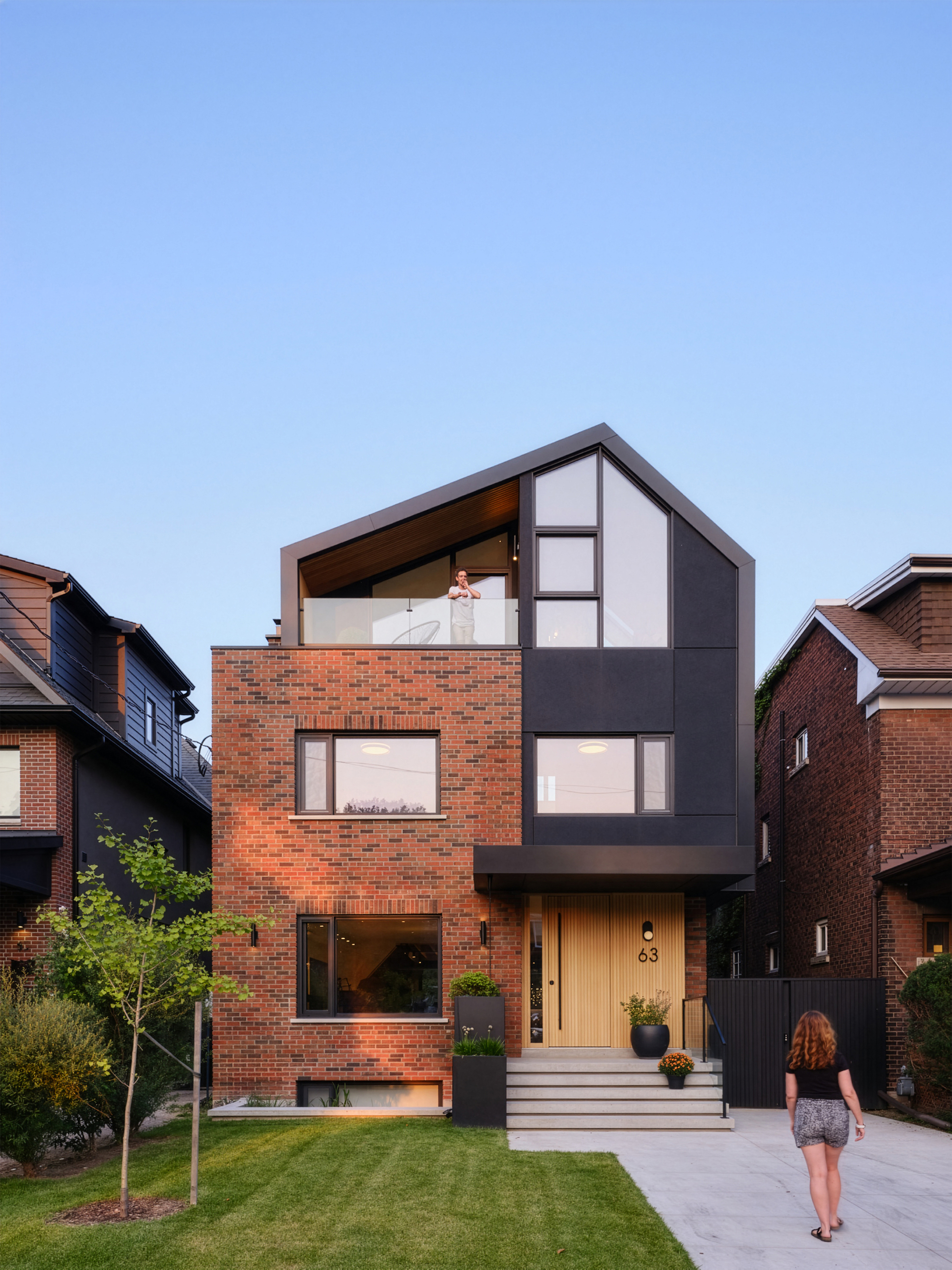 Street facade of brick and black cladding with a gabled top floor, a person on the balcony and a woman walking past at dusk