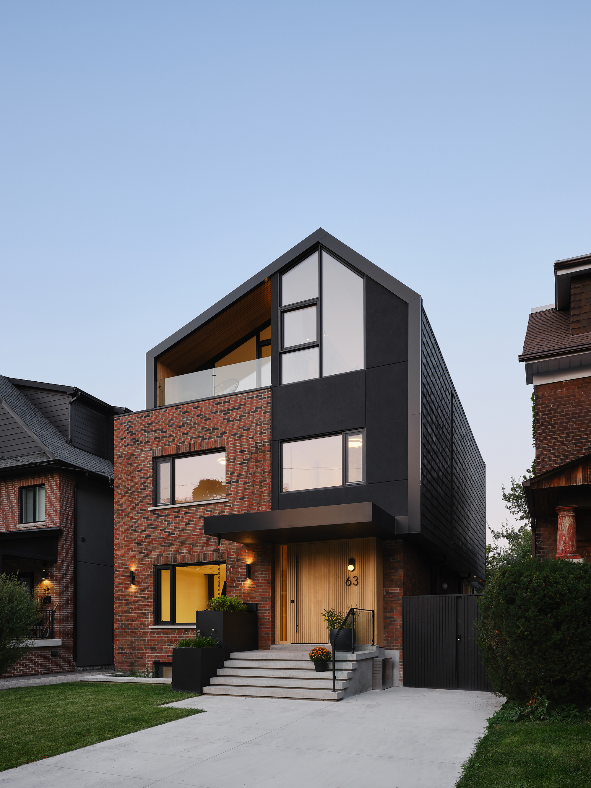 Front elevation of the brick and black-clad house with a wood entry door and glowing windows under a clear evening sky