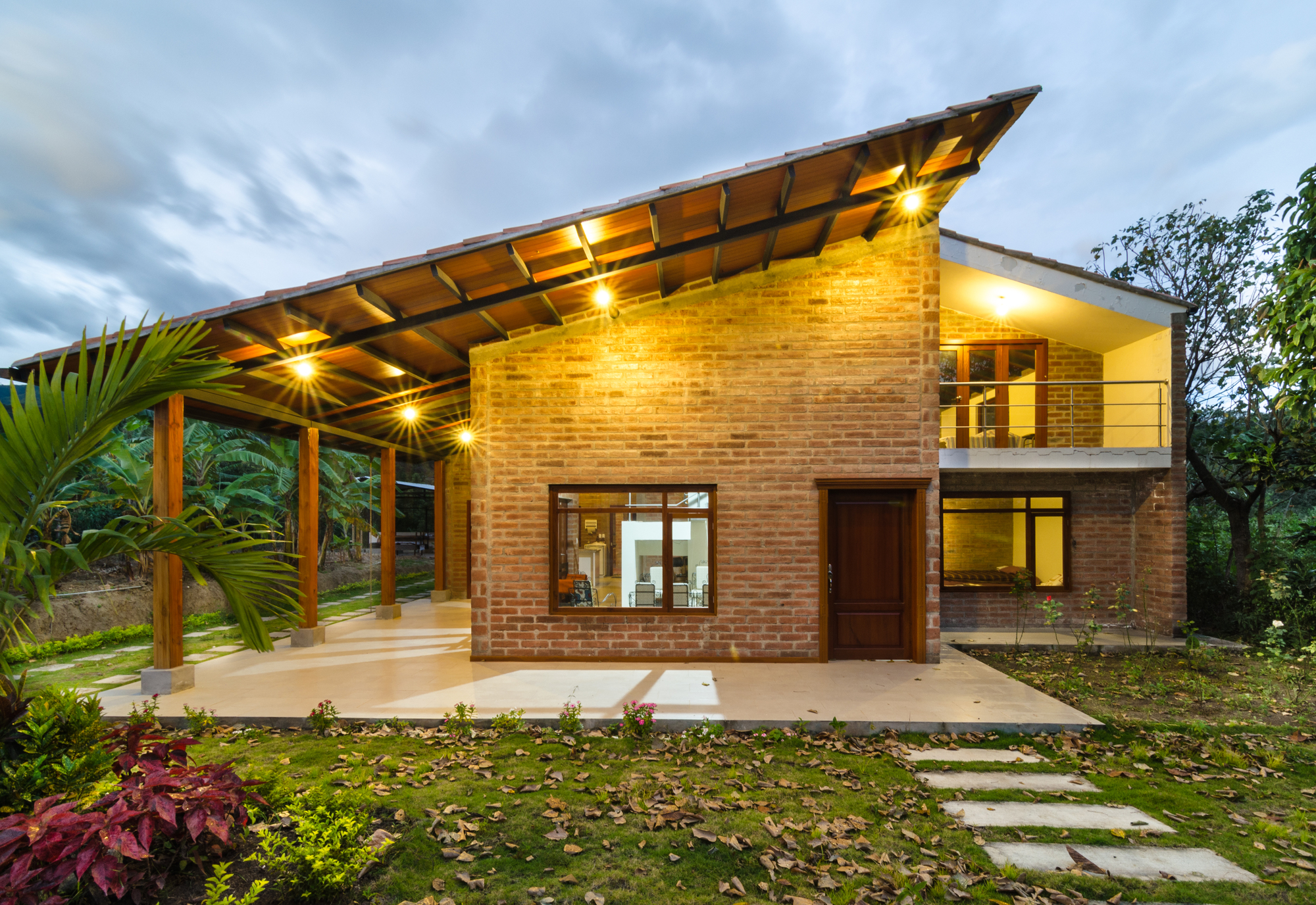Brick house facade under a steeply pitched timber roof with warm downlights, seen at dusk beside a lawn with stepping stones