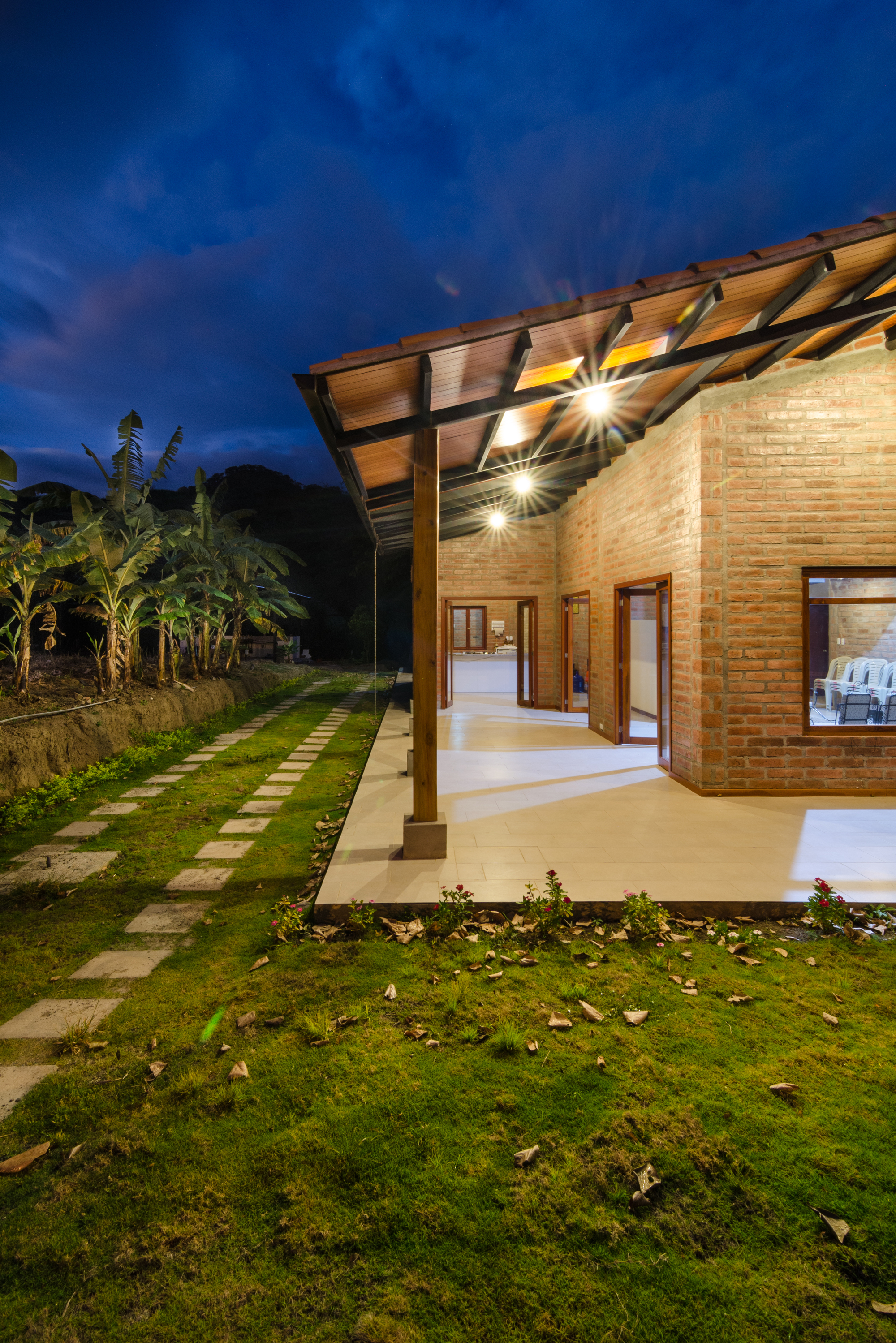 Covered porch with a timber column, exposed beams and glowing ceiling lights, framed by banana plants at night