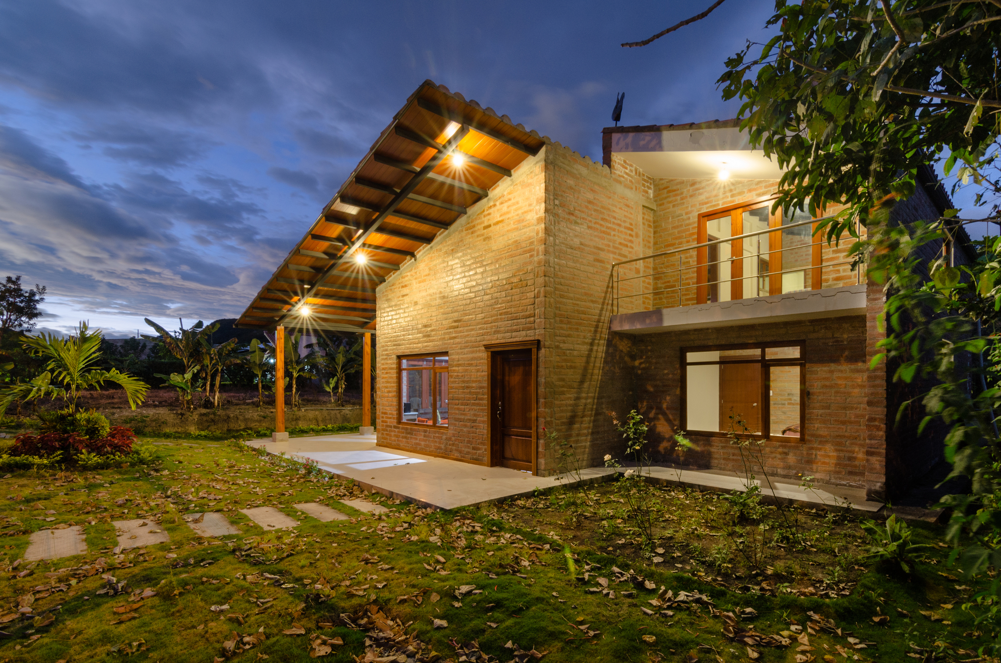 Corner of the two-storey brick house with a sloping timber roof and balcony railing, lit against a blue evening sky