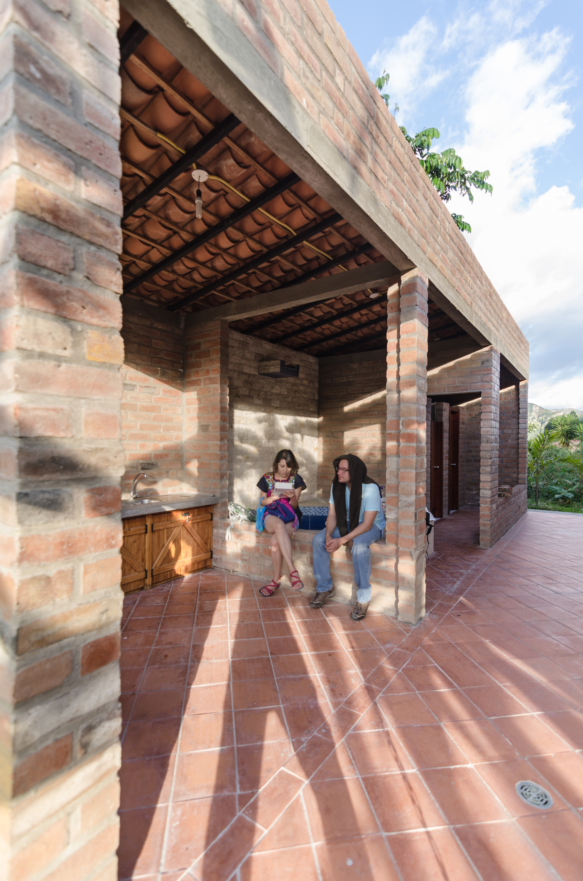 Open brick porch with terracotta floor tiles and exposed tile roof, where two people sit talking in afternoon shadows