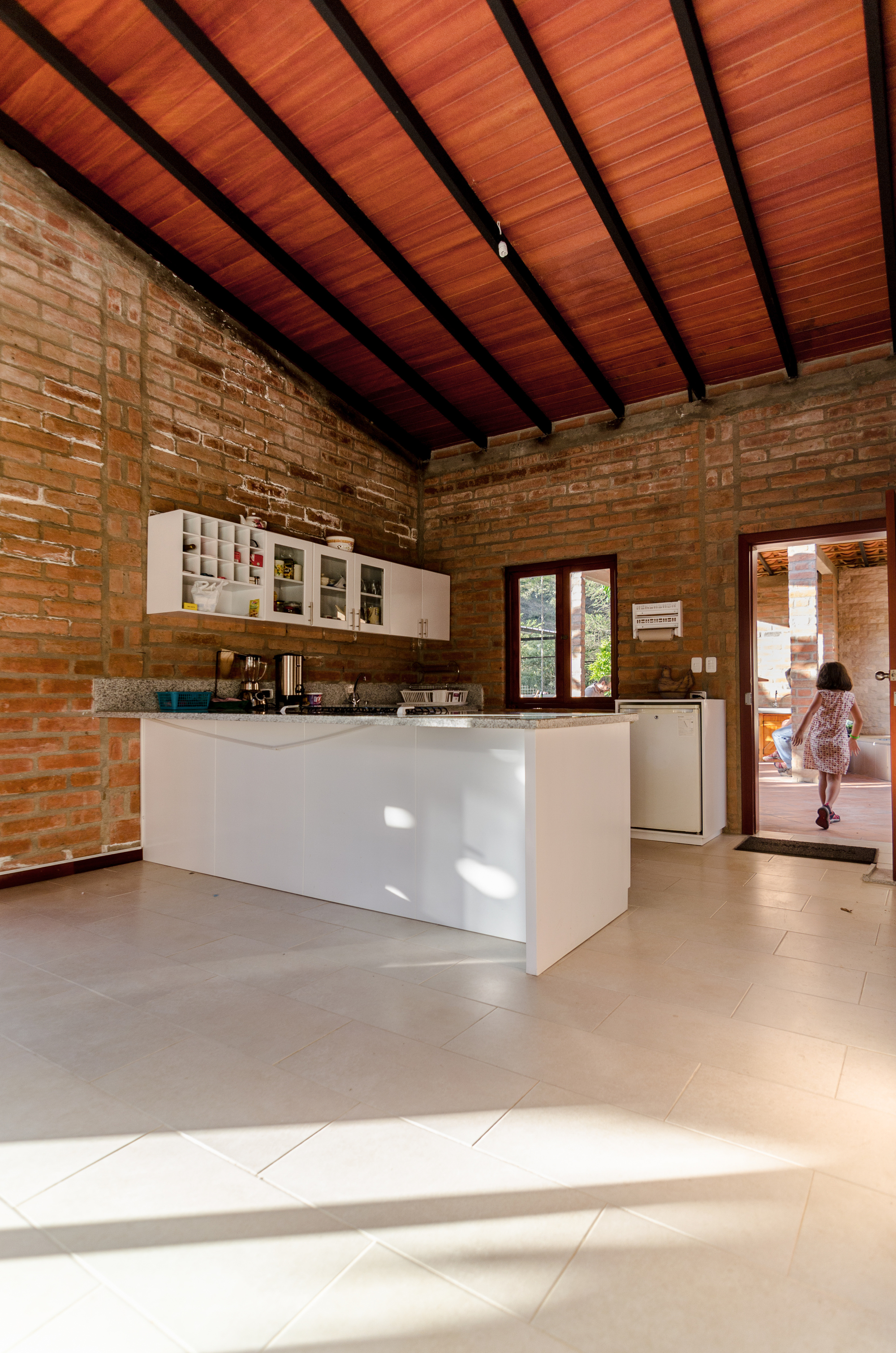 Kitchen with white island and wall cabinets against exposed brick, under a timber ceiling, with a child walking through the sunlit doorway