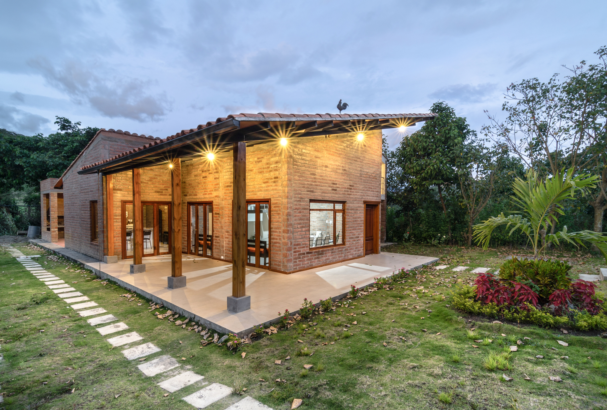Brick house with timber porch columns and glowing downlights at dusk, a rooster figure on the roof and stepping stones across the lawn