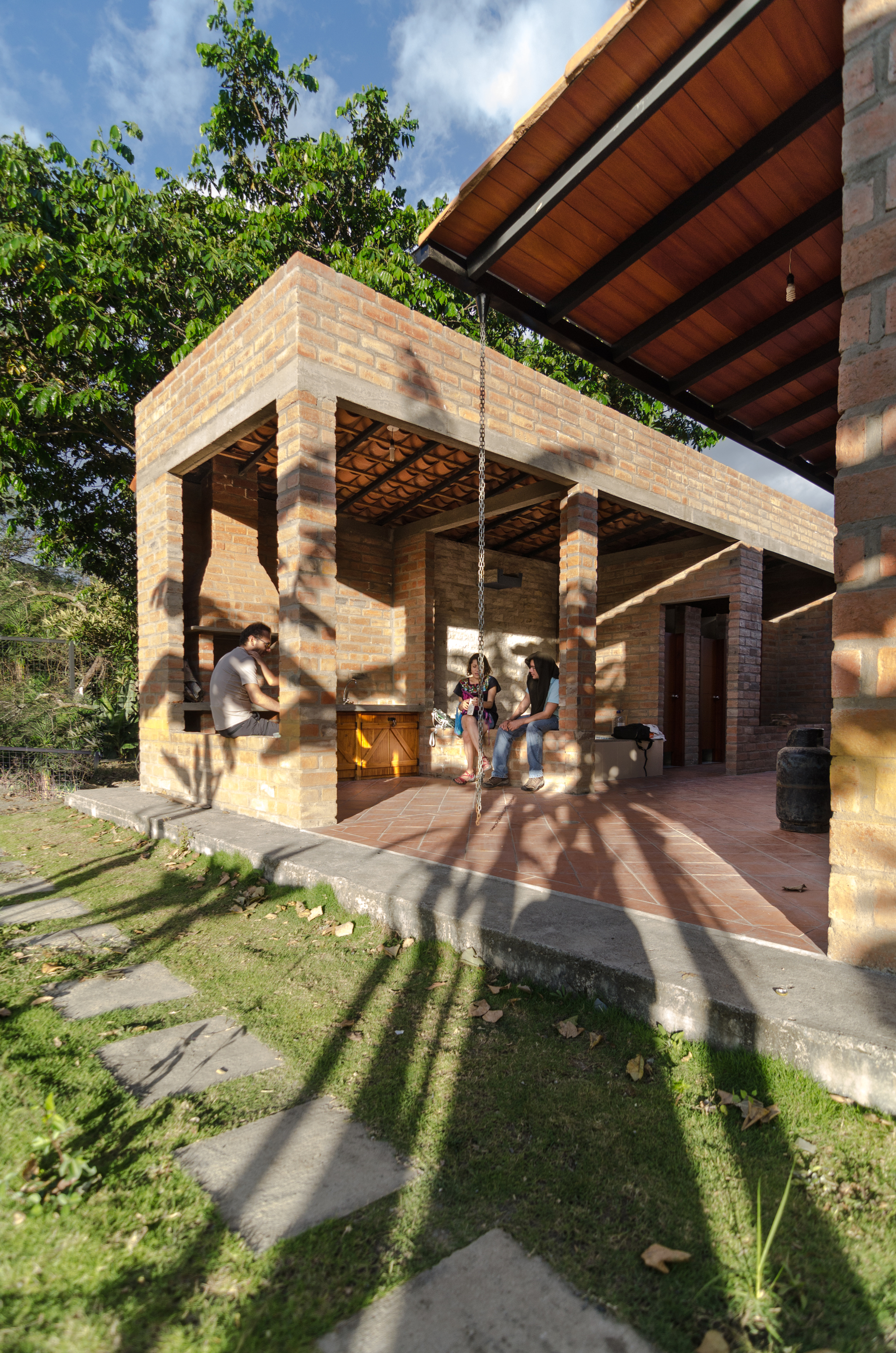 Open brick pavilion with tiled floor where people sit in the shade, tree shadows falling across the lawn and stepping stones