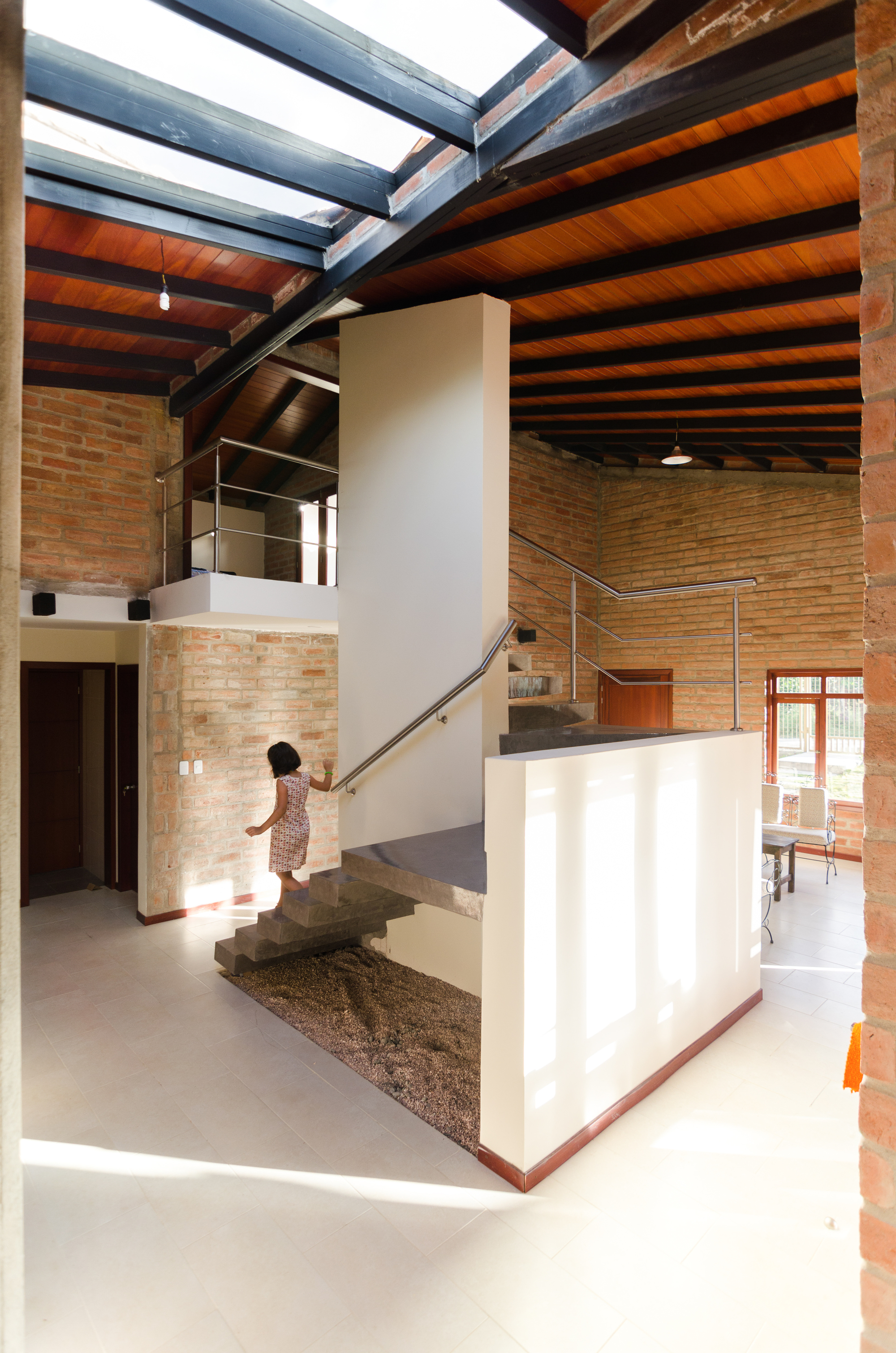 Double-height living space with a concrete stair over a gravel bed, skylight beams overhead and a woman walking down in daylight