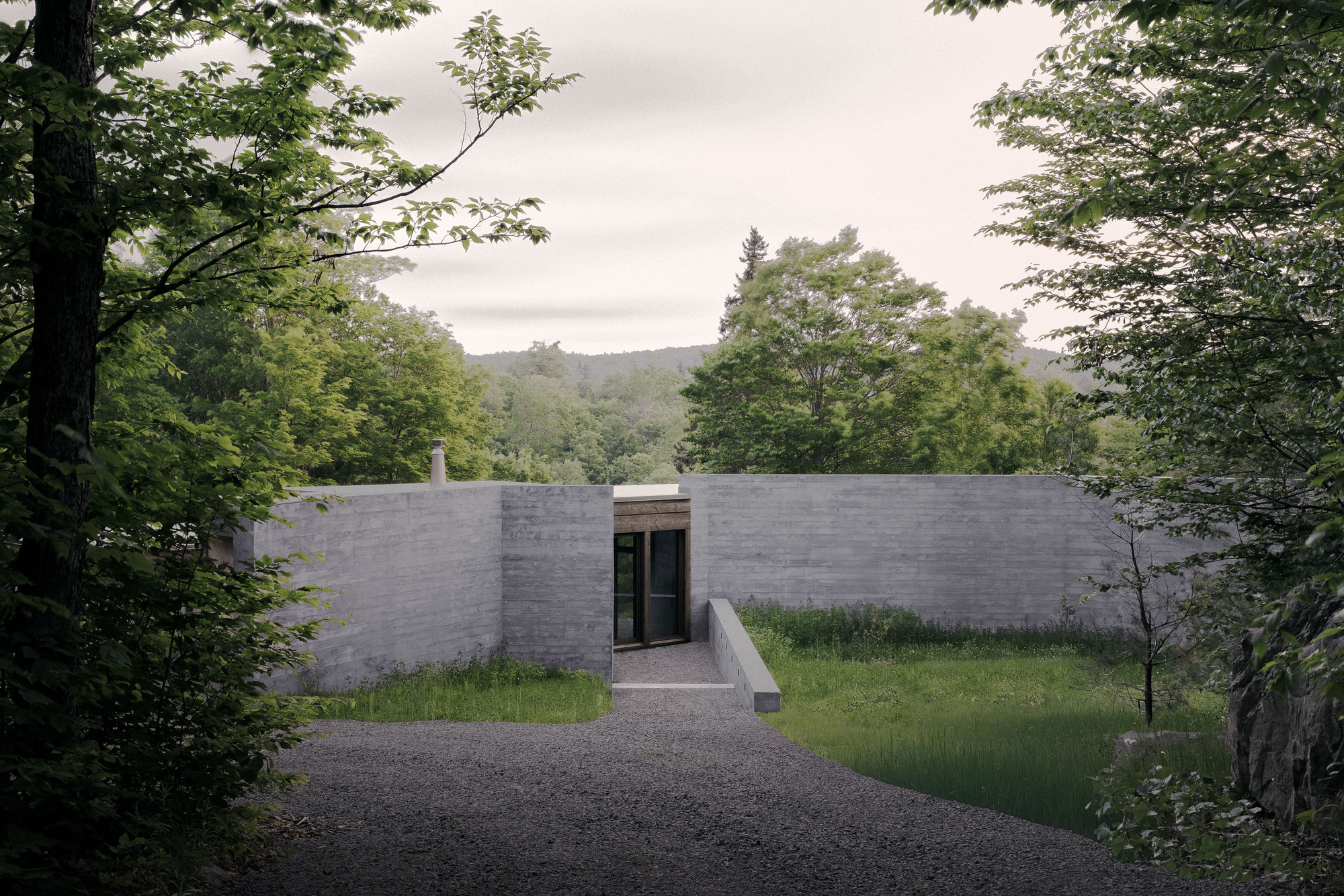 Board-formed concrete walls flanking a recessed glazed entry, reached by a gravel drive through summer woodland under an overcast sky